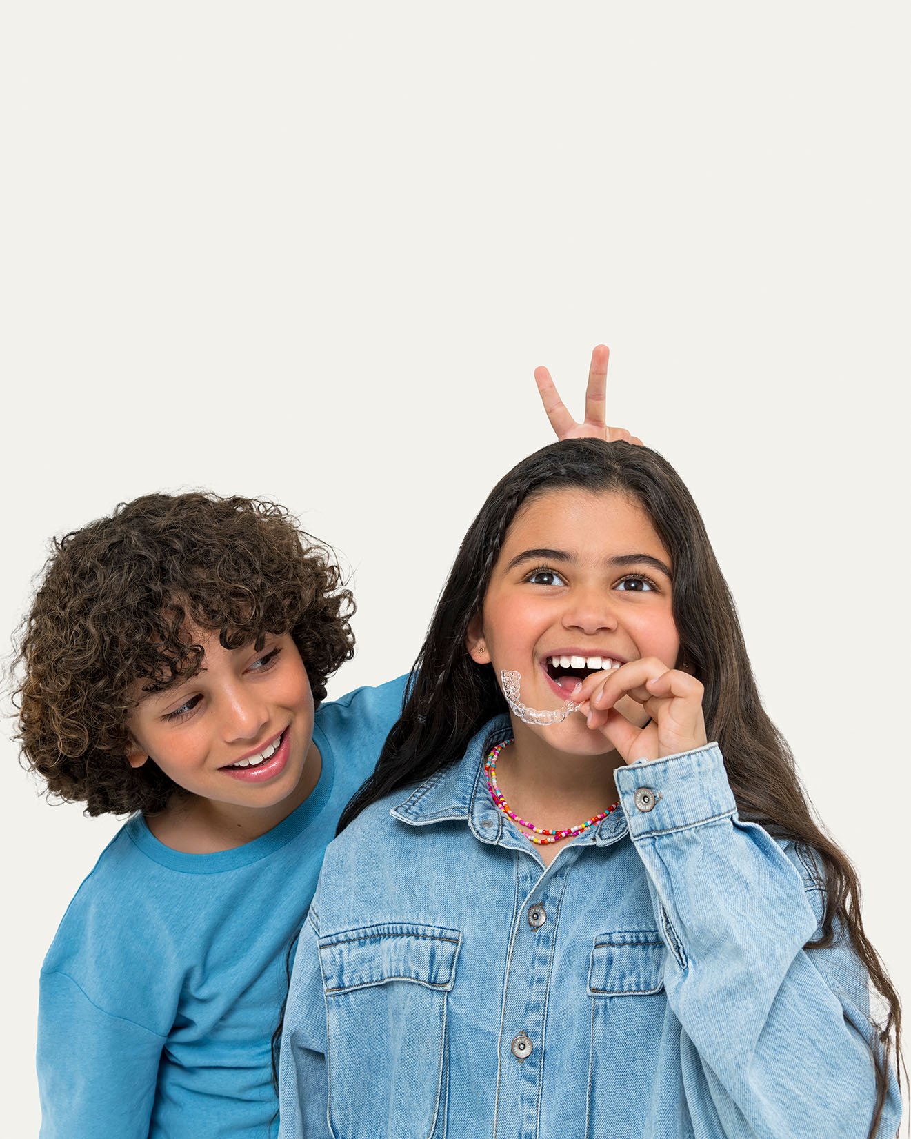 Two kids, a boy with curly hair and a girl with straight hair wearing a denim jacket, playing and smiling against a plain background, with the girl making bunny ears behind the boy.