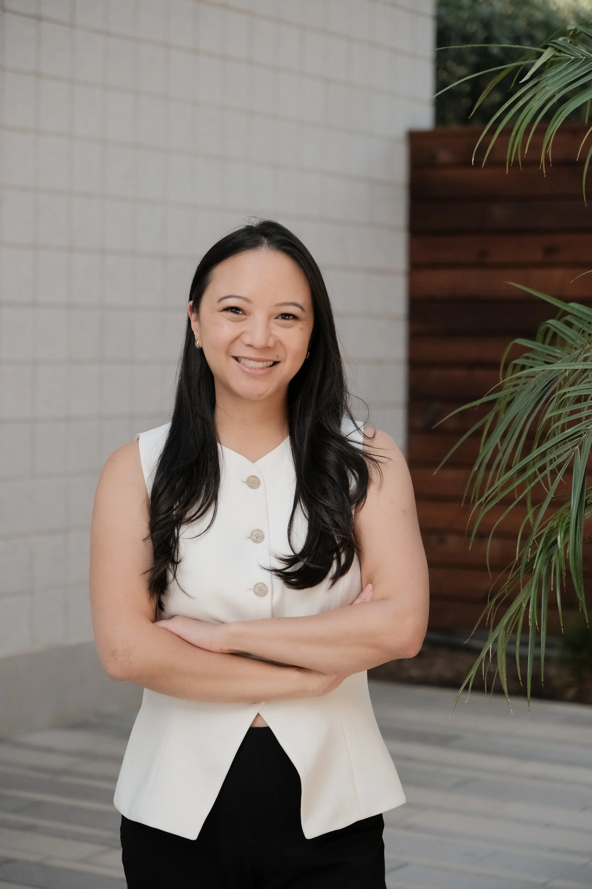 A smiling woman with long dark hair, wearing a sleeveless white top with buttons, standing with her arms crossed in front of a background of a white brick wall and wooden fence, with green plants visible on the right side.