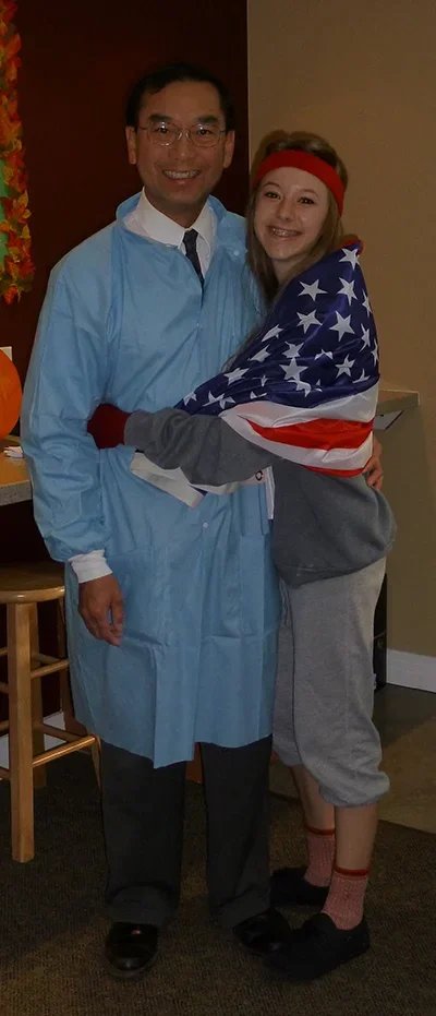 A man dressed as a nurse and a young woman wrapped in an American flag hugging each other indoors.