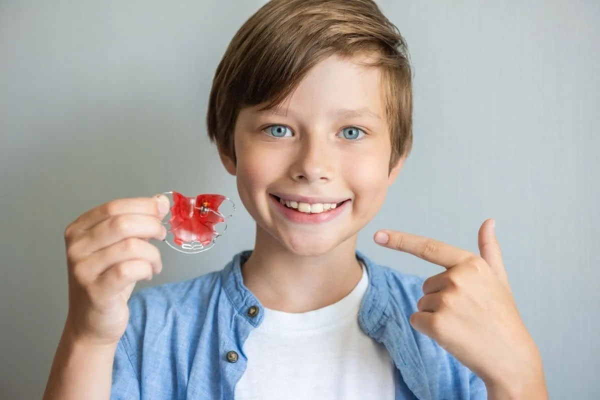 A young boy with blue eyes and brown hair smiling and pointing to a red dental retainer he is holding with his right hand.