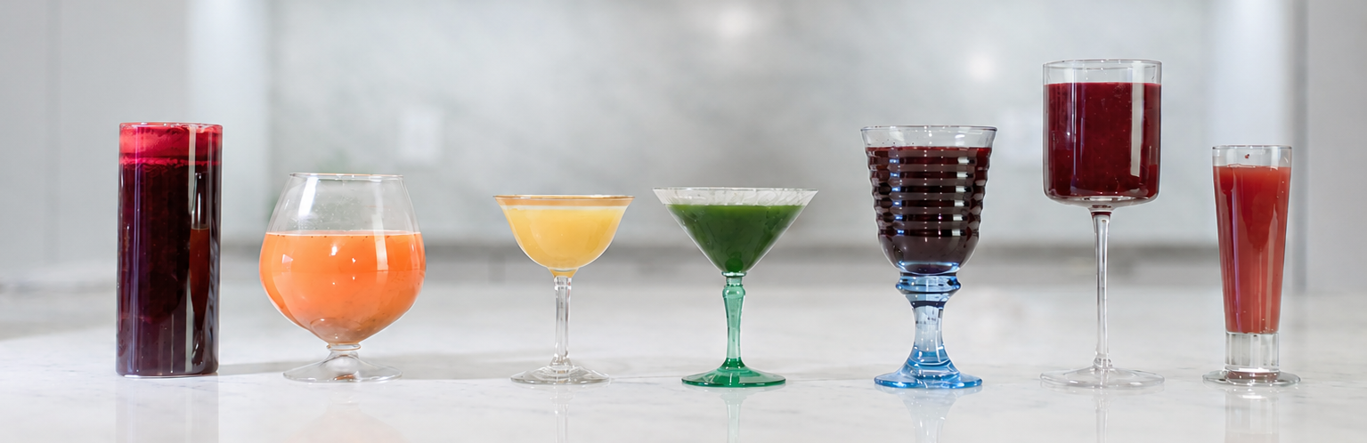 lineup of 7 beautiful juices in different glasses, backdrop a clean white marble kitchen, from left to right red, orange, yellow, green, blue, purple, pink