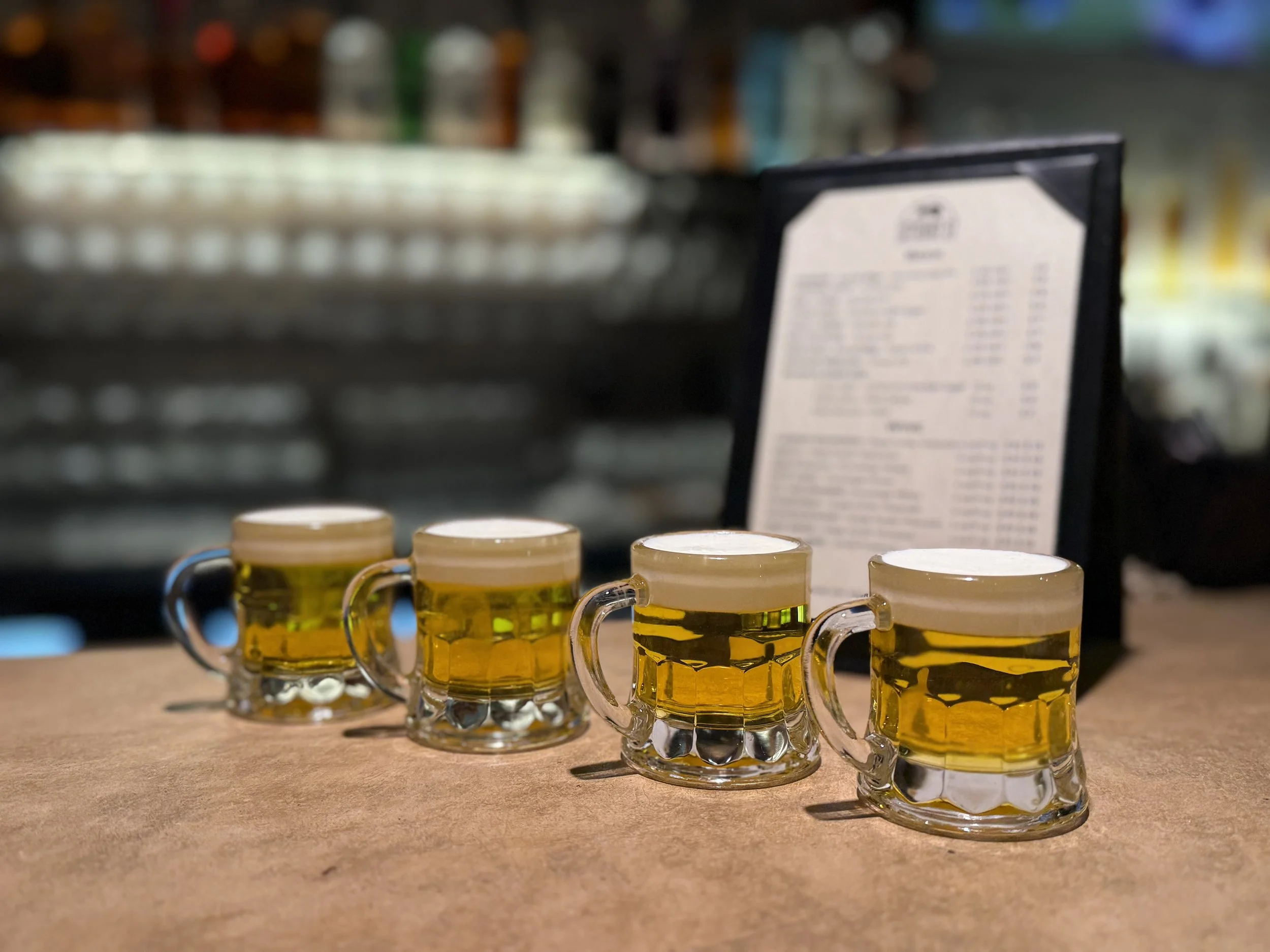 Four small glass mugs filled with beer on a counter with a blurred menu in the background.