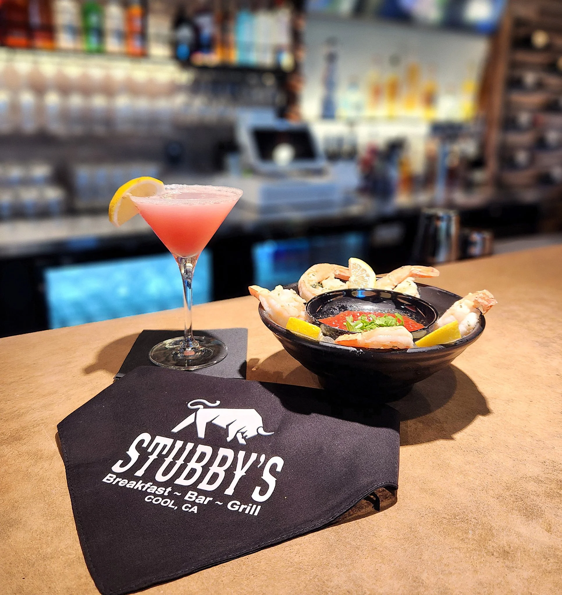 A pink cocktail with a lemon wedge garnish, a black bowl of shrimp tempura with dipping sauce, and a black napkin with the logo of Stubby's restaurant in Clear Lake, California, on a wooden bar counter.