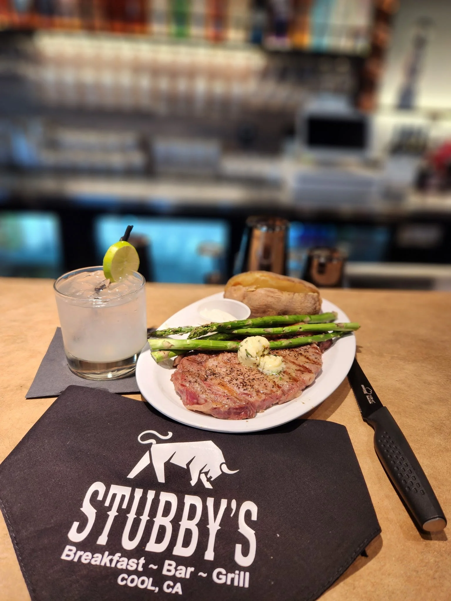 A plate of cooked steak served with asparagus, a baked potato with sour cream, and a small cup of butter, accompanied by a cocktail with lime wedge, on a table at Stubby's breakfast, bar, and grill in Cool, California.