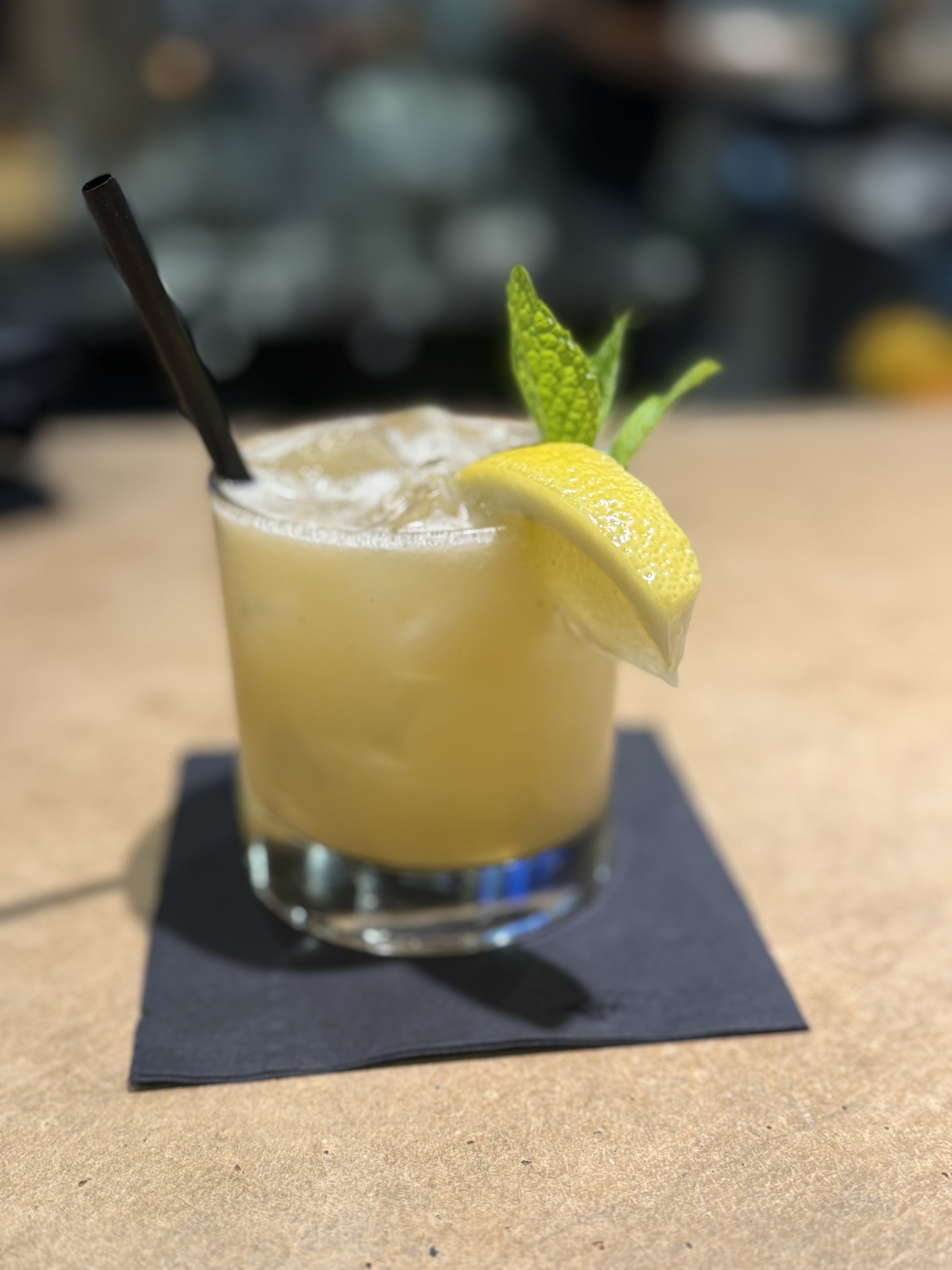 A yellow cocktail garnished with a lemon wedge and mint leaves, with a black straw, on a black napkin on a wooden surface.
