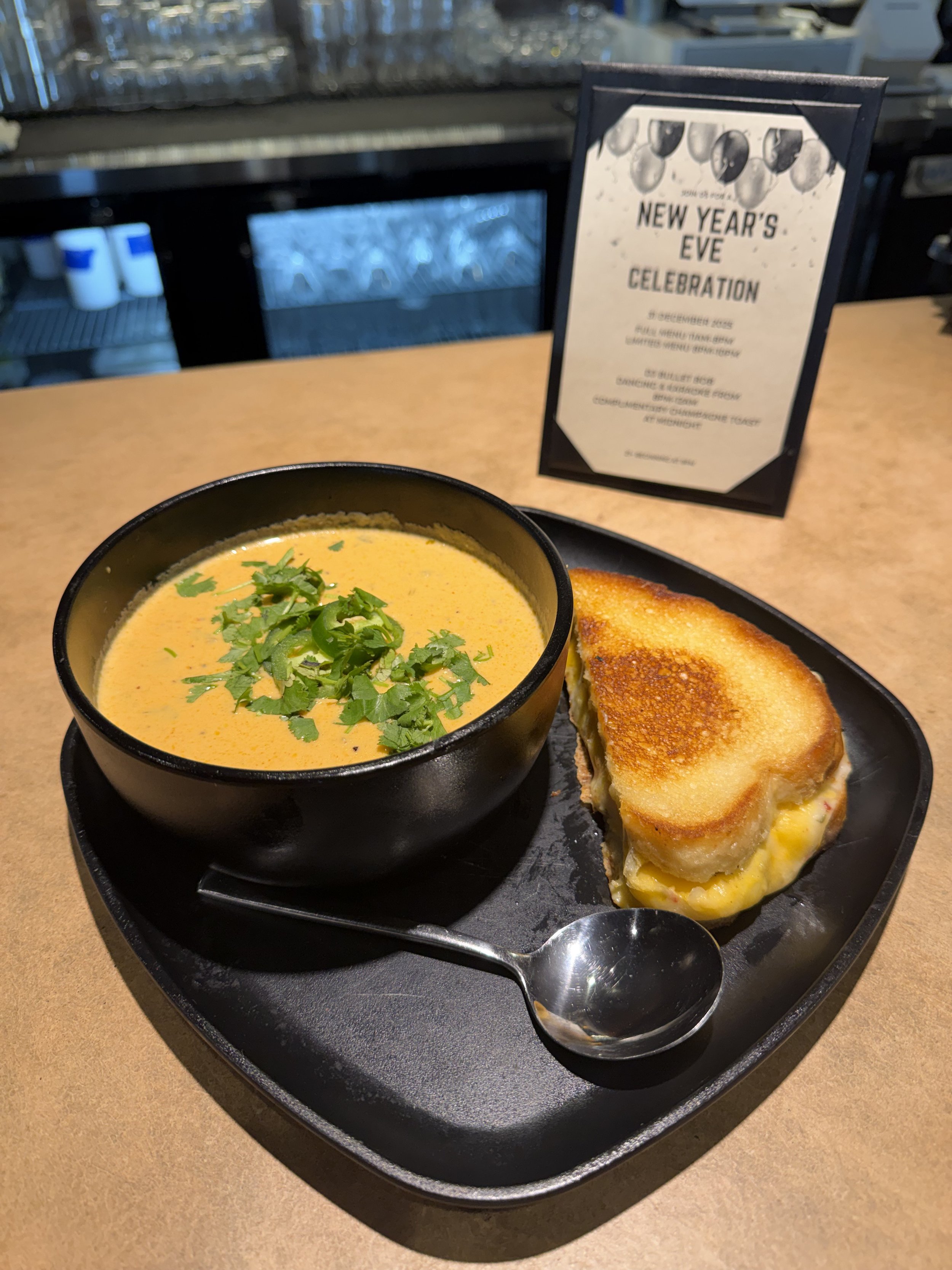 A bowl of creamy yellow soup garnished with chopped cilantro, served with a toasted grilled cheese sandwich on a black tray.