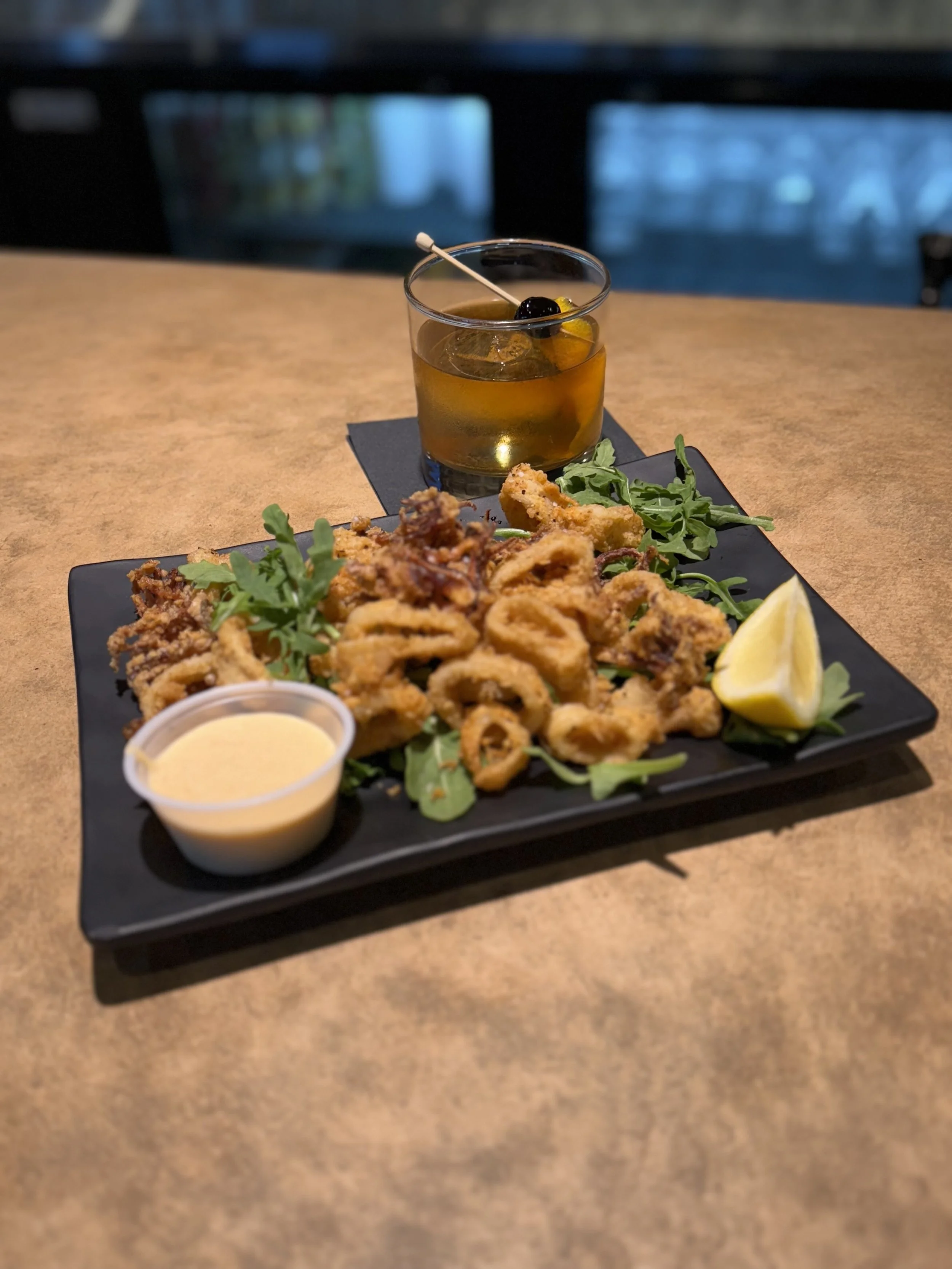 Fried calamari served with a lemon wedge, a side dipping sauce, arugula garnish, and a cocktail with a cherry and lemon slice