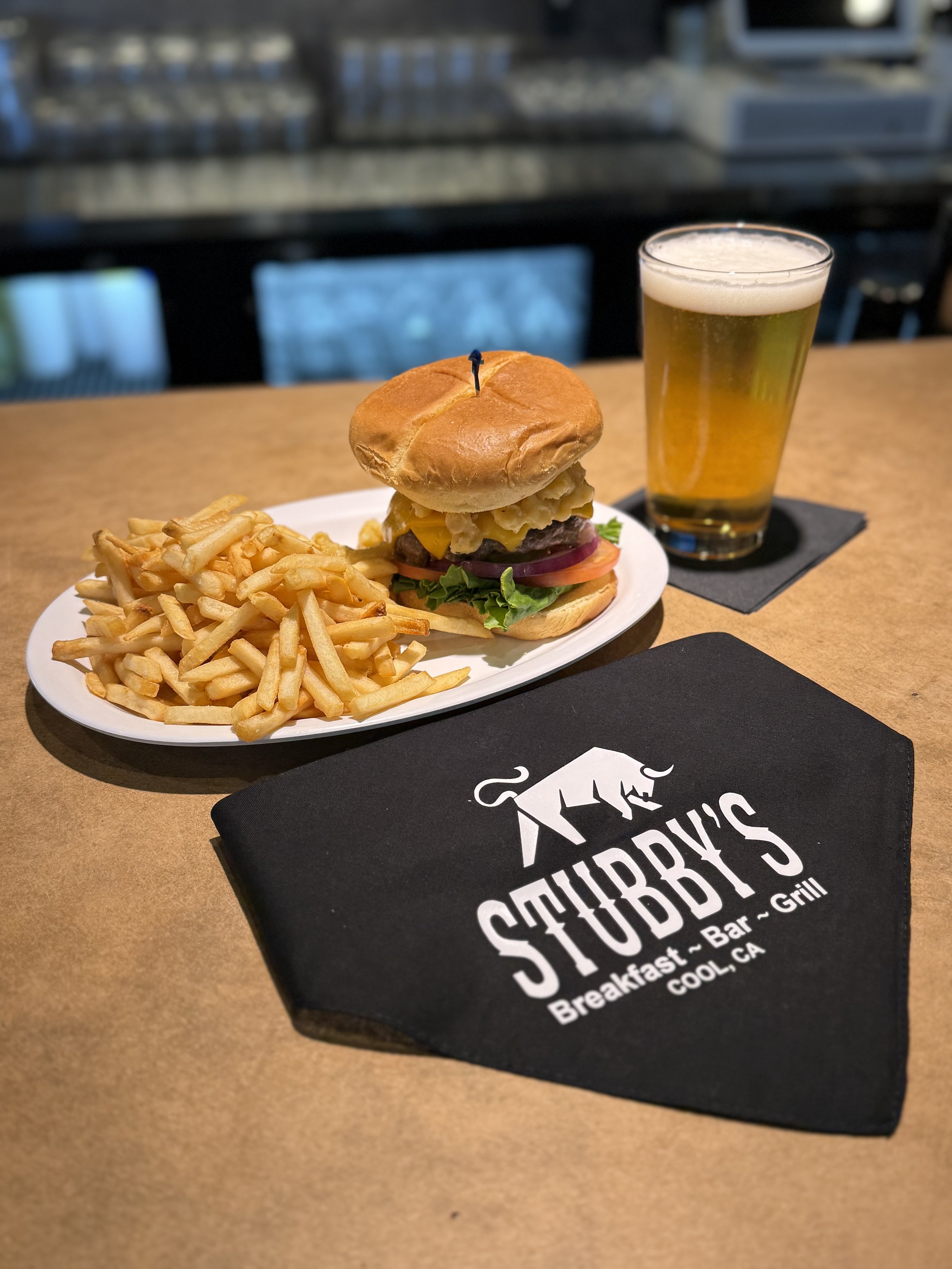 A burger with lettuce, tomato, cheese, and a fried onion ring on a bun, served with French fries and a glass of beer on a table at Stubb's in Cool, California.