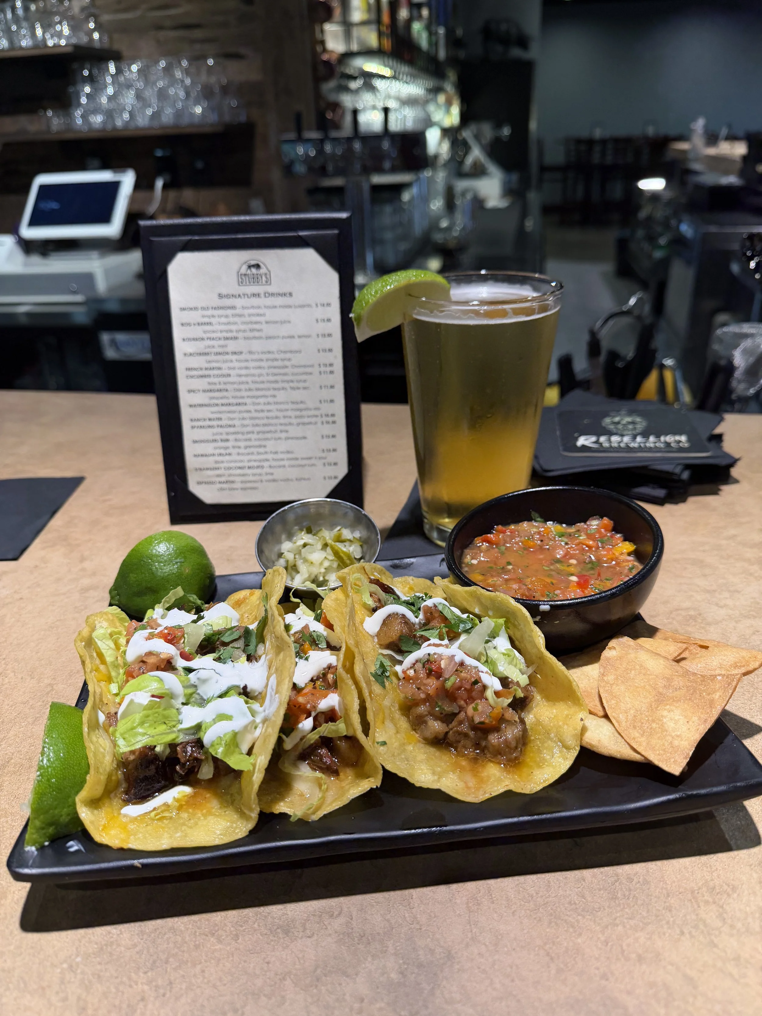 A plate of three tacos with diced beef, chopped lettuce, sour cream, and cilantro garnished with lime wedges, alongside a small bowl of salsa, a lemon-lime soda in a tall glass with a lime wedge, and tortilla chips, all set on a black tray at a resta