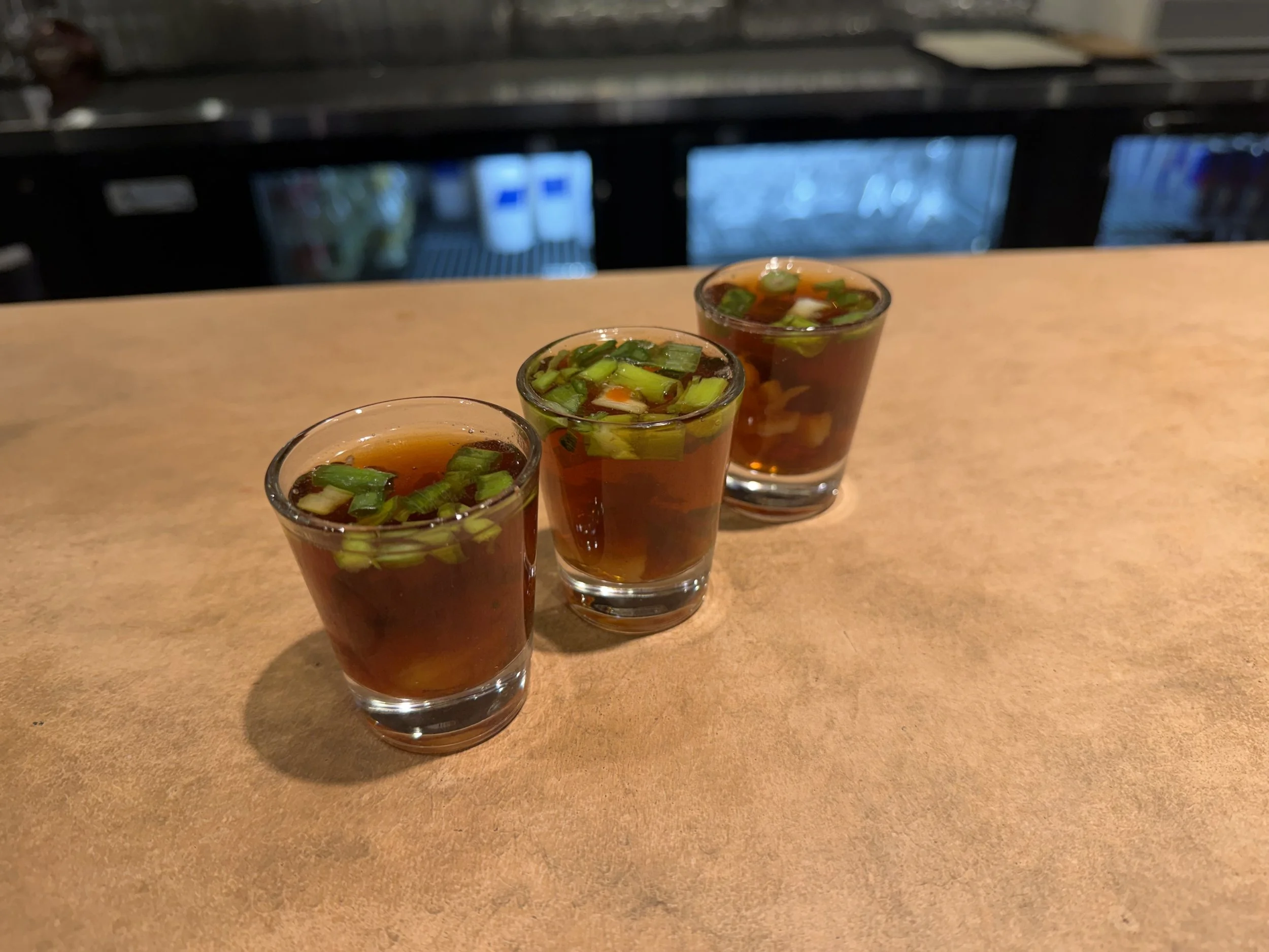 Three small shot glasses filled with a dark liquid and garnished with chopped green onions, lined up on a wooden surface.