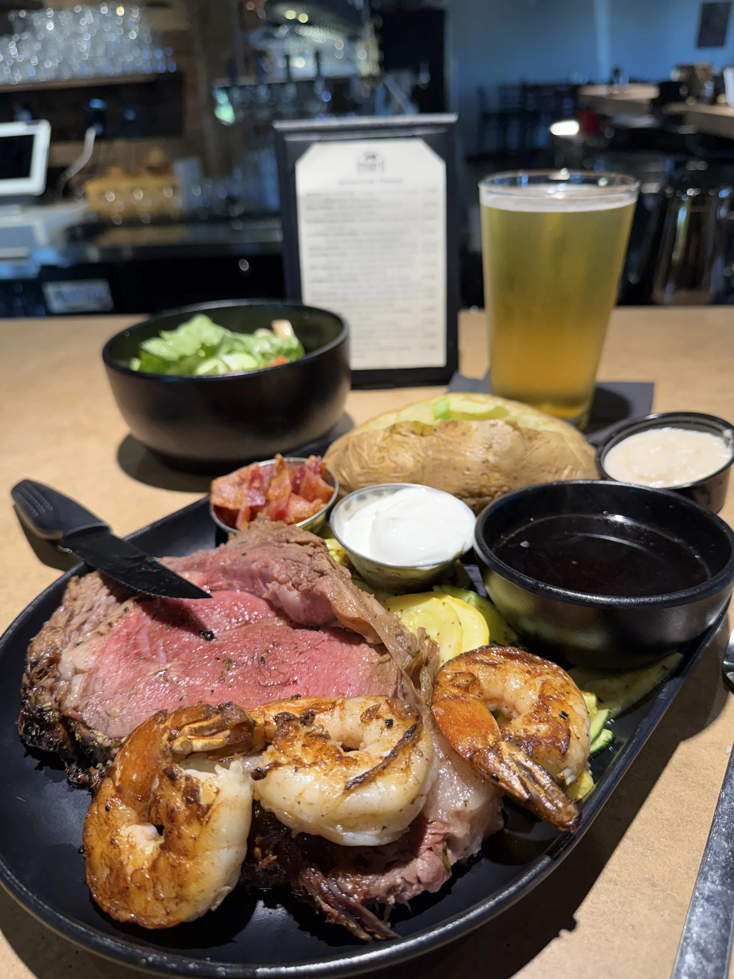A surf and turf plate with a grilled steak, shrimp, baked potato, salads, sauces, and a beverage, served at a restaurant.