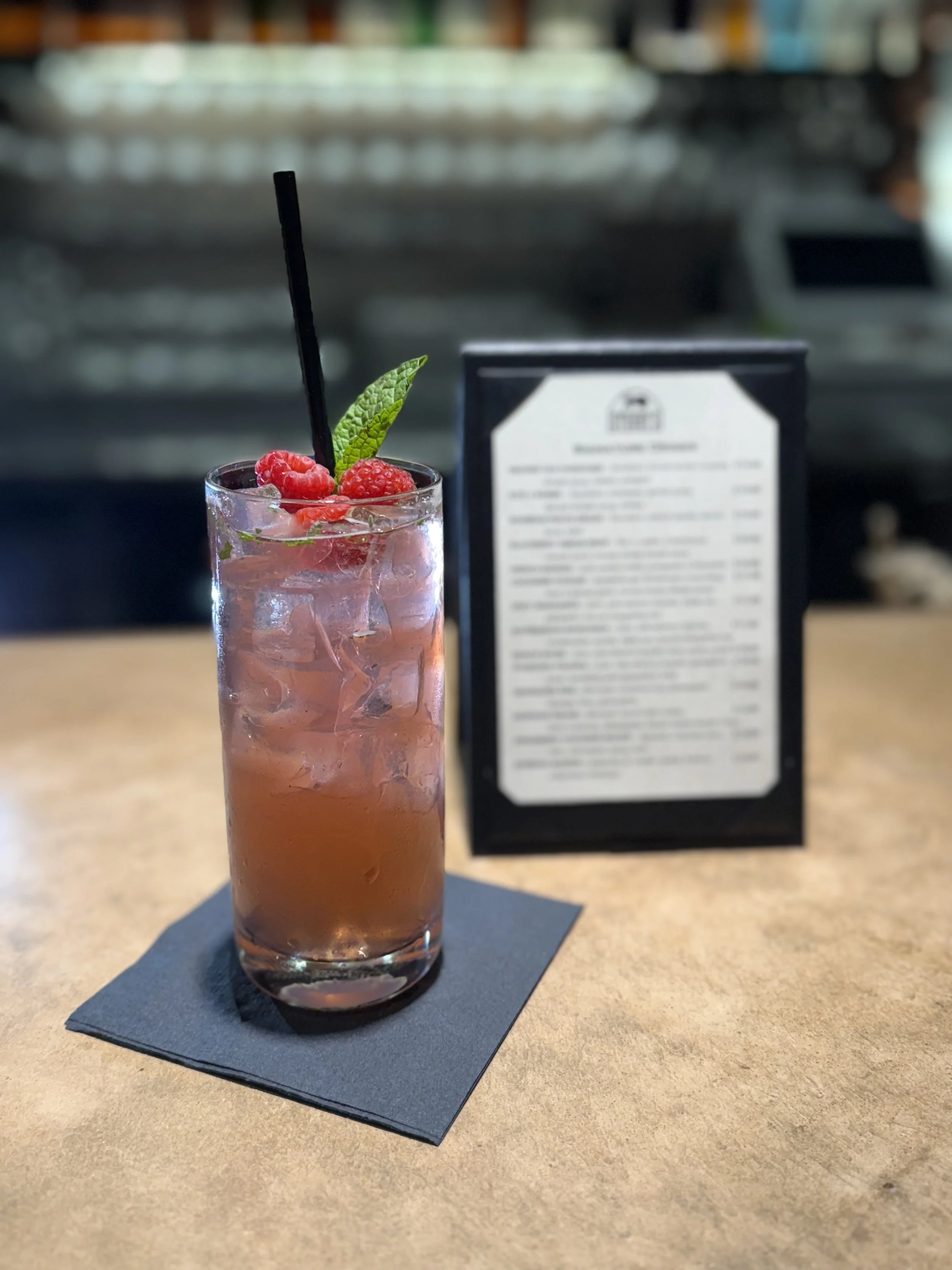 A tall glass of pinkish cocktail garnished with fresh raspberries and a green mint leaf, served with a black straw on a black napkin on a wooden table, with a blurred menu and bar counter in the background.