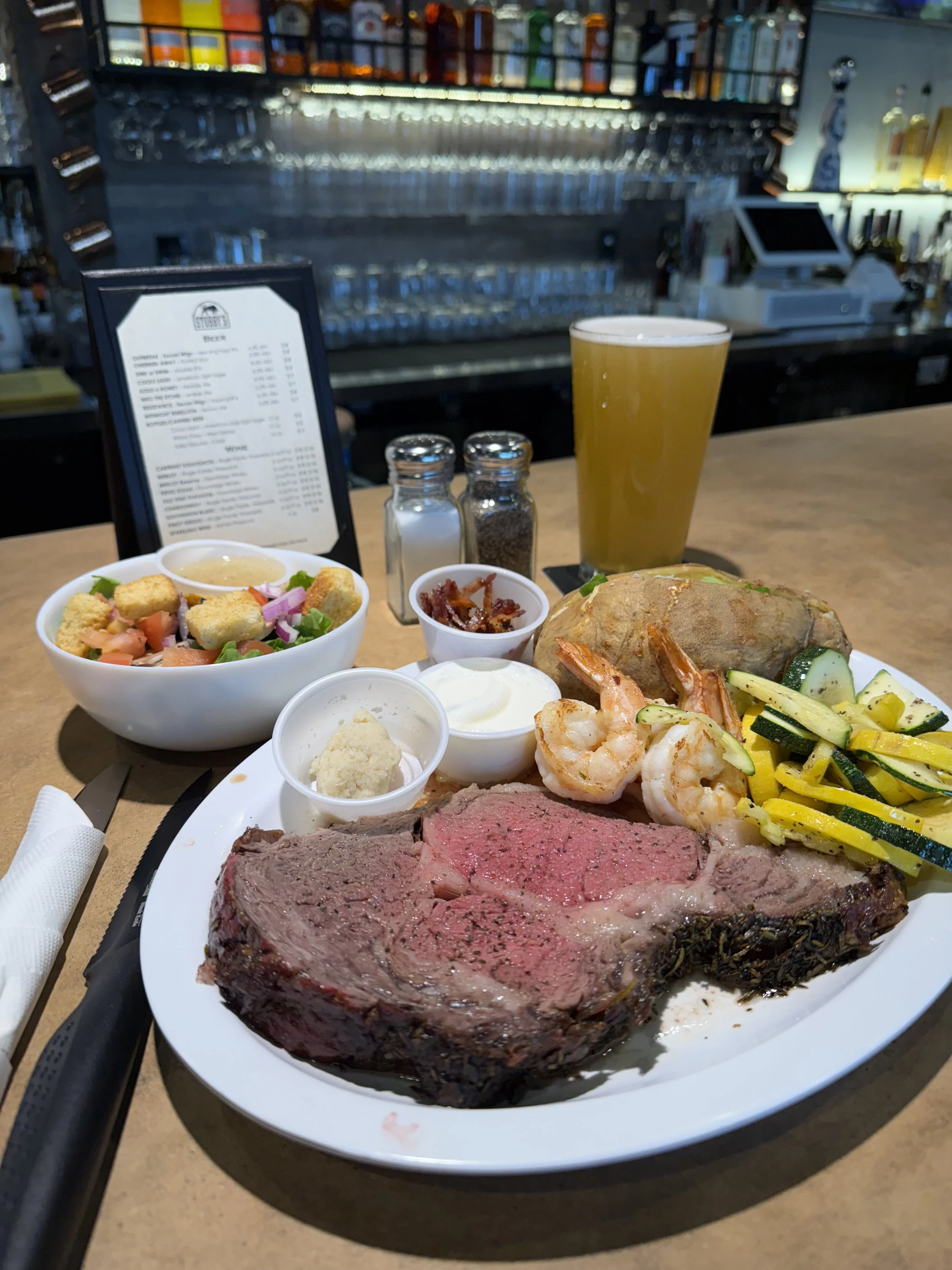 A dinner plate with a large cut of medium-rare steak, shrimp, grilled vegetables, and a baked potato. Side dishes include a salad, sour cream, butter, and a small bowl of what appears to be sun-dried tomatoes or bacon bits. There is also a glass of b