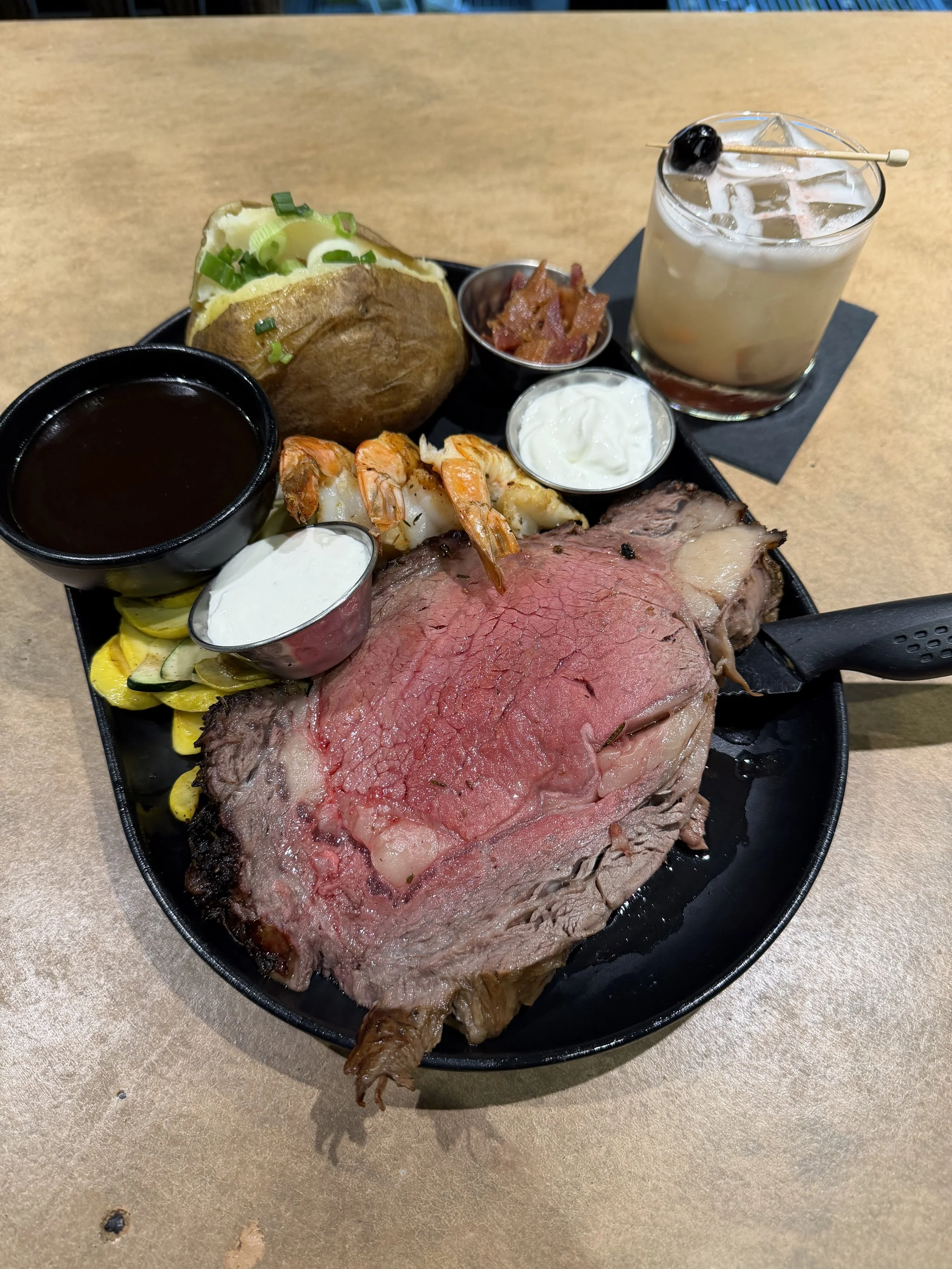 A steak dinner platter with a large cut of rare beef, shrimp, baked potato topped with green onions, and various sides including sauces, bacon, and vegetables, accompanied by a cocktail garnished with an olive.