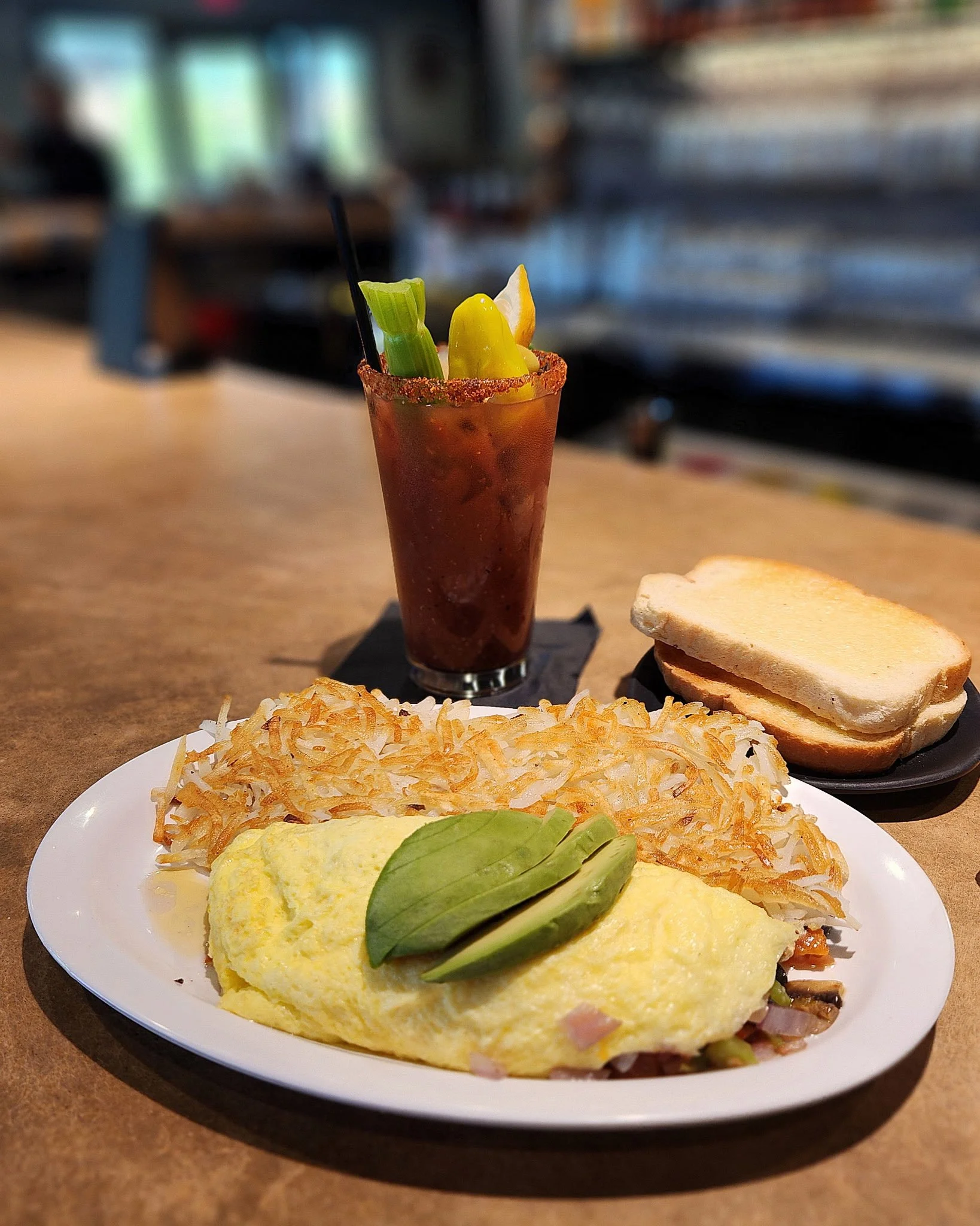 A breakfast plate with an omelet garnished with basil, hash browns, and toast, with a Bloody Mary cocktail topped with celery and lemon on a bar or restaurant table.