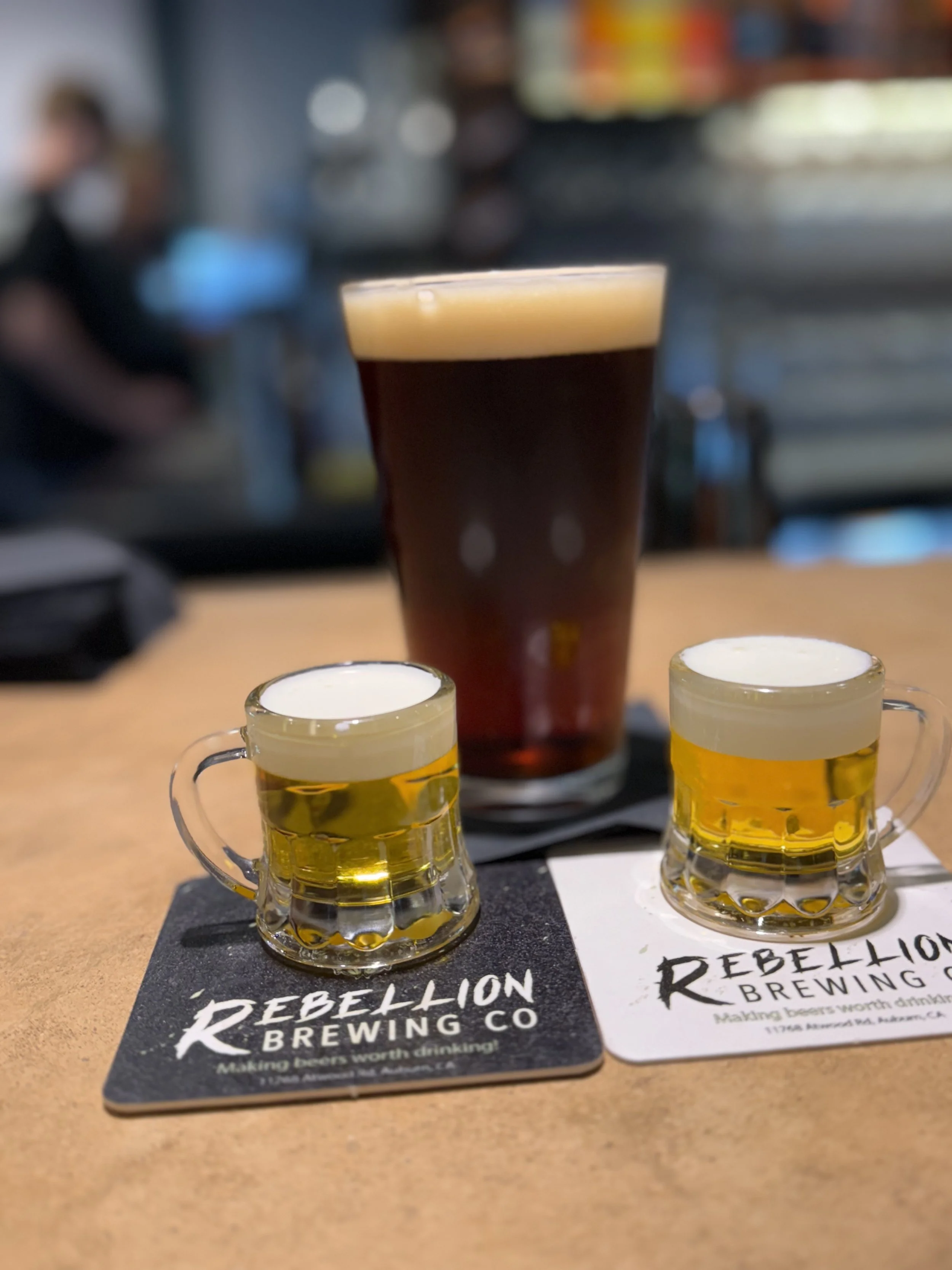 A pint glass of dark beer and two small glass mugs of light beer on coasters from Rebellion Brewing Co. on a wooden table in a bar with blurred background.