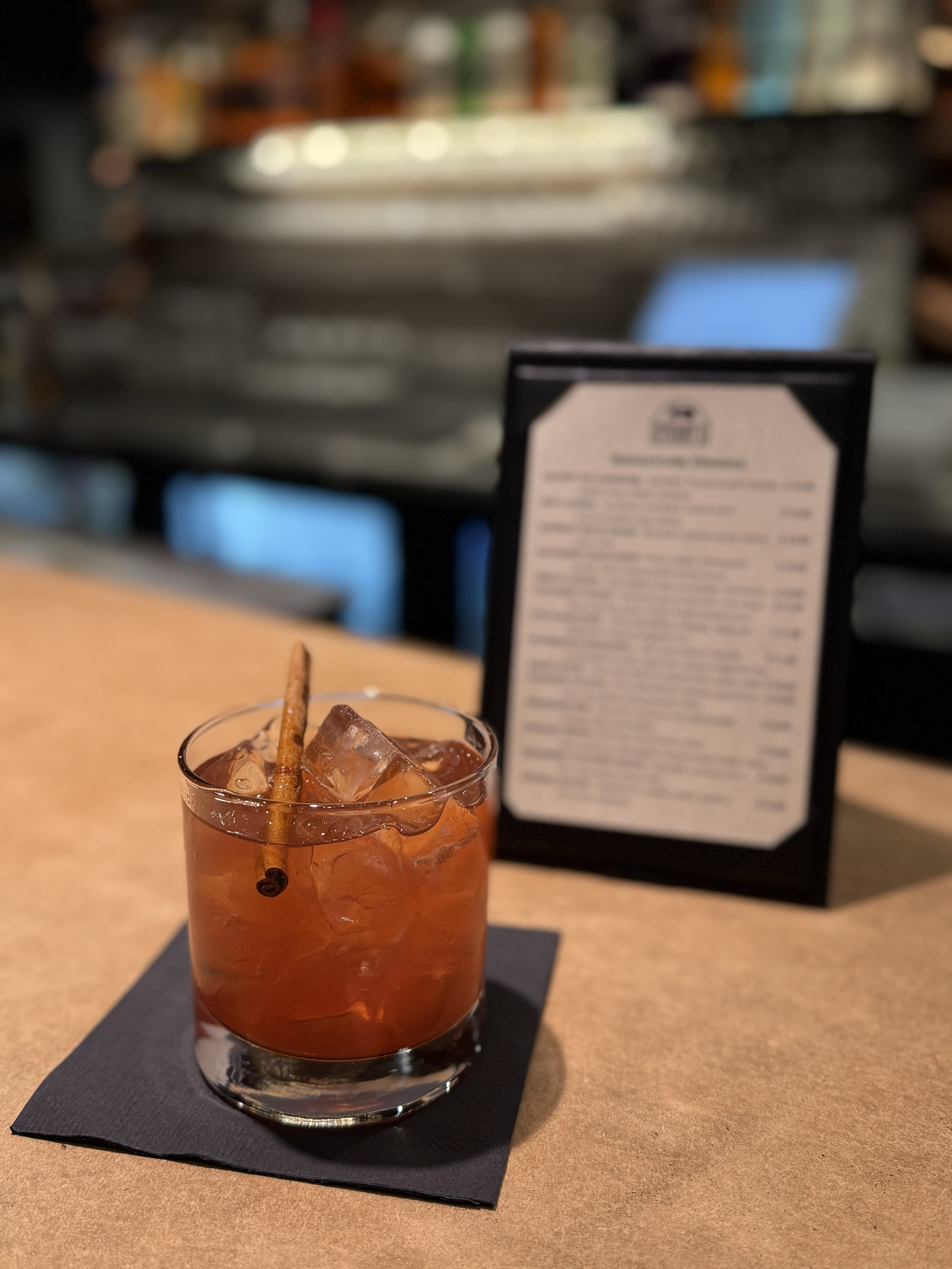 An old-fashioned cocktail with ice and a cinnamon stick garnished in a short glass on a black napkin at a bar or restaurant, with a blurred menu in the background.