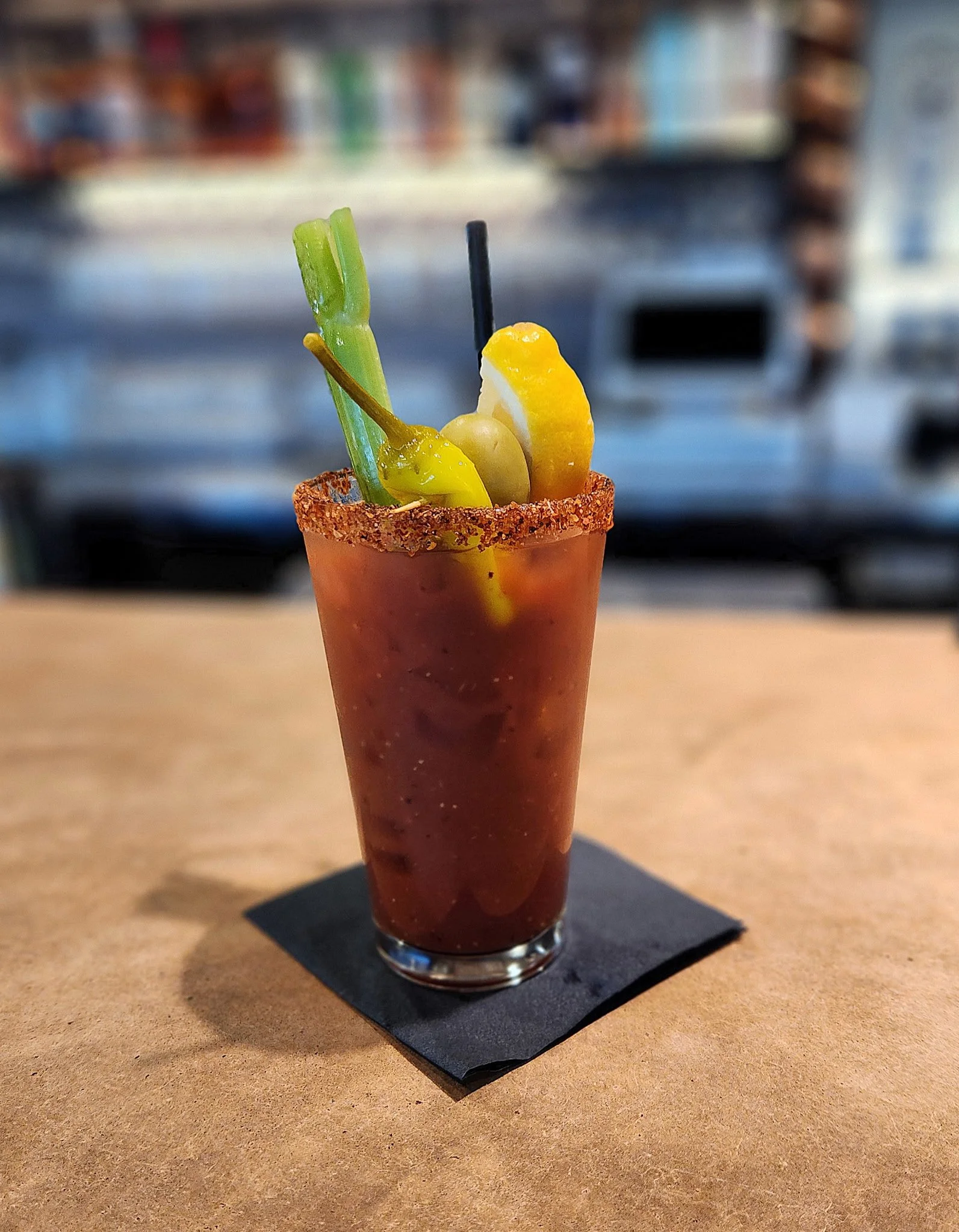 A Bloody Mary cocktail garnished with celery, lemon, green bean, and green Olive, with a salted rim, on a black napkin on a wooden table.