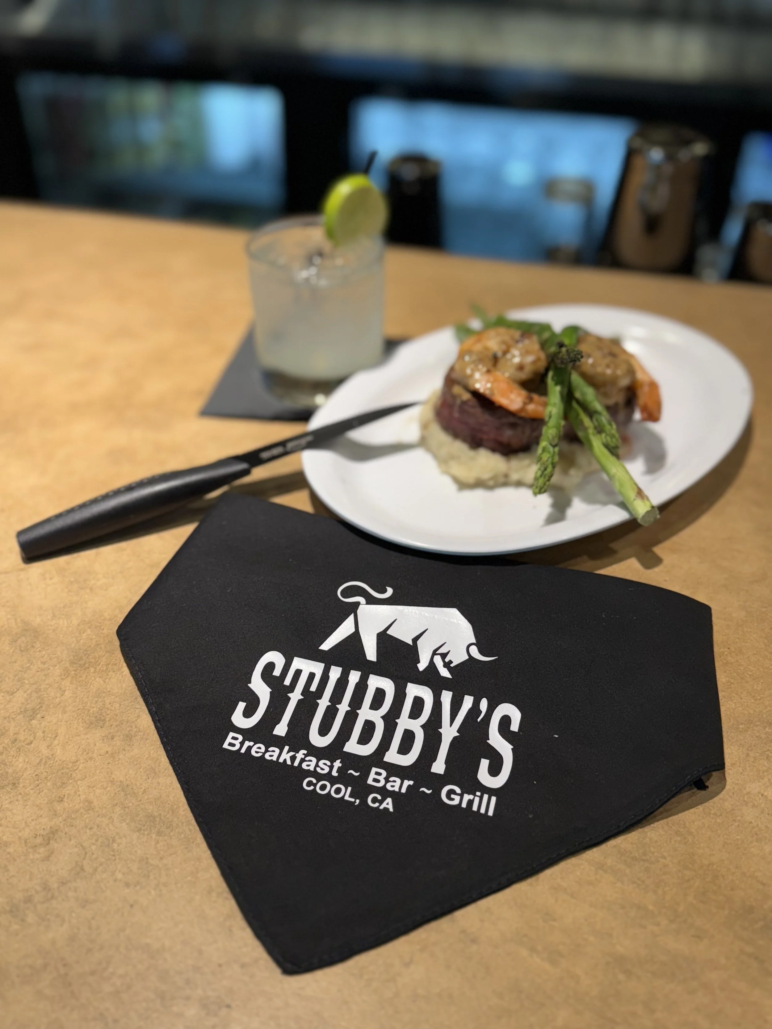 A plate with a filet mignon topped with shrimp, served with asparagus on a bed of mashed potatoes. A drink with a lime wedge is in the background, along with a black Stubby's napkin on a wooden table.