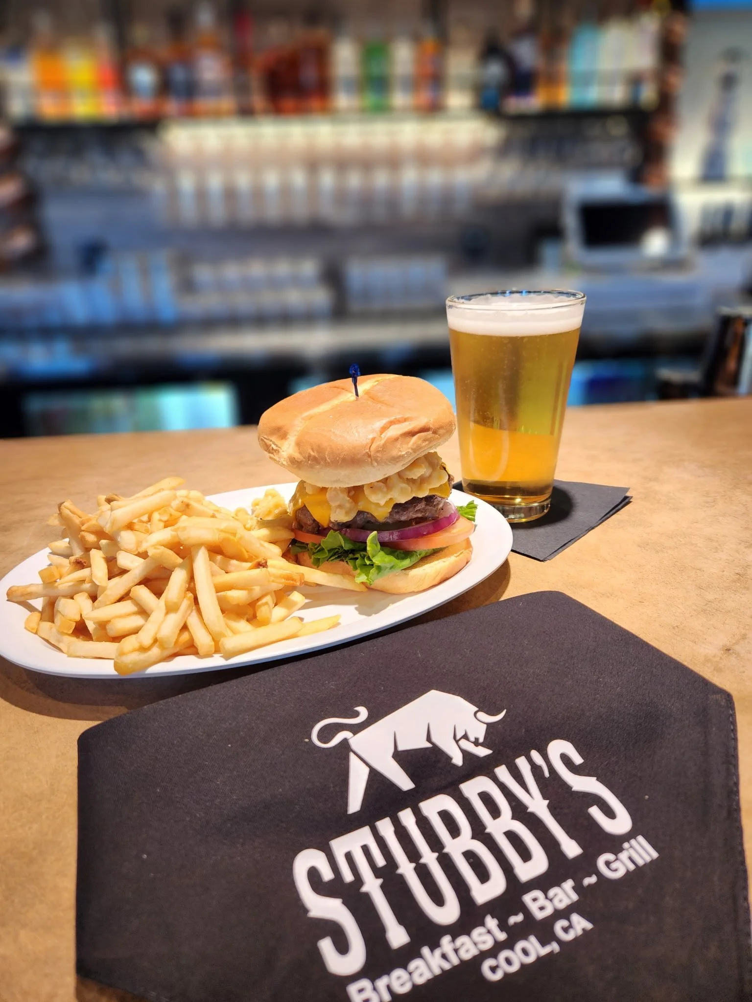 A plate with French fries and a burger with lettuce, red onions, yellow cheese, and scrambled eggs on a bun, accompanied by a glass of beer on a bar counter with a Stubb's restaurant napkin in the foreground.
