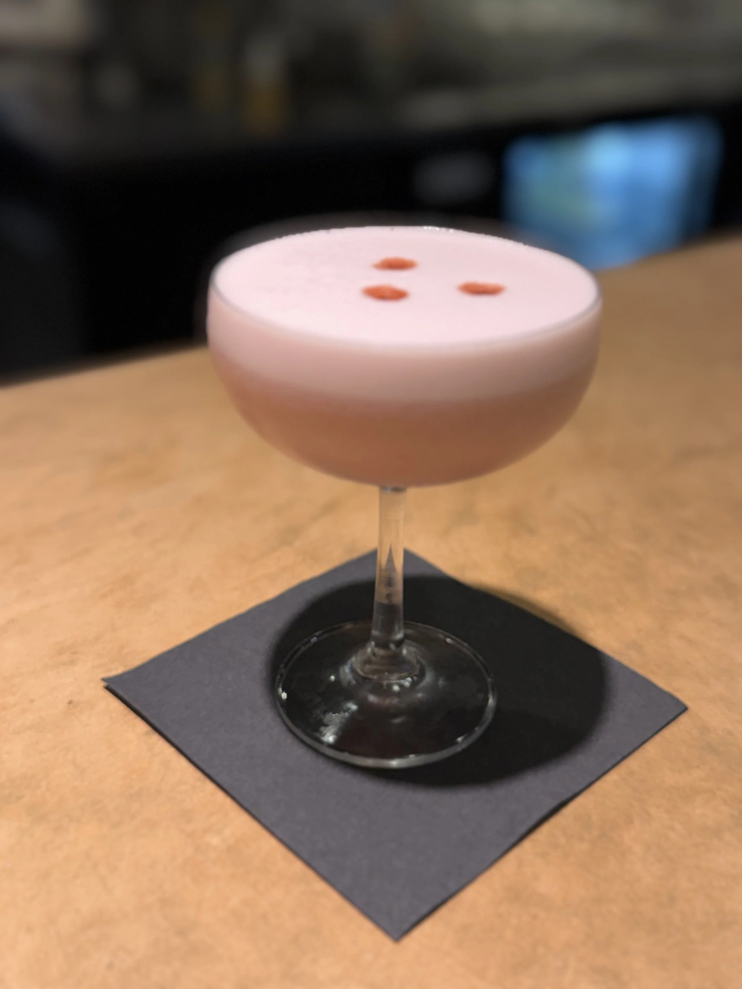 A pink cocktail with orange dots on top in a coupe glass on a black napkin.