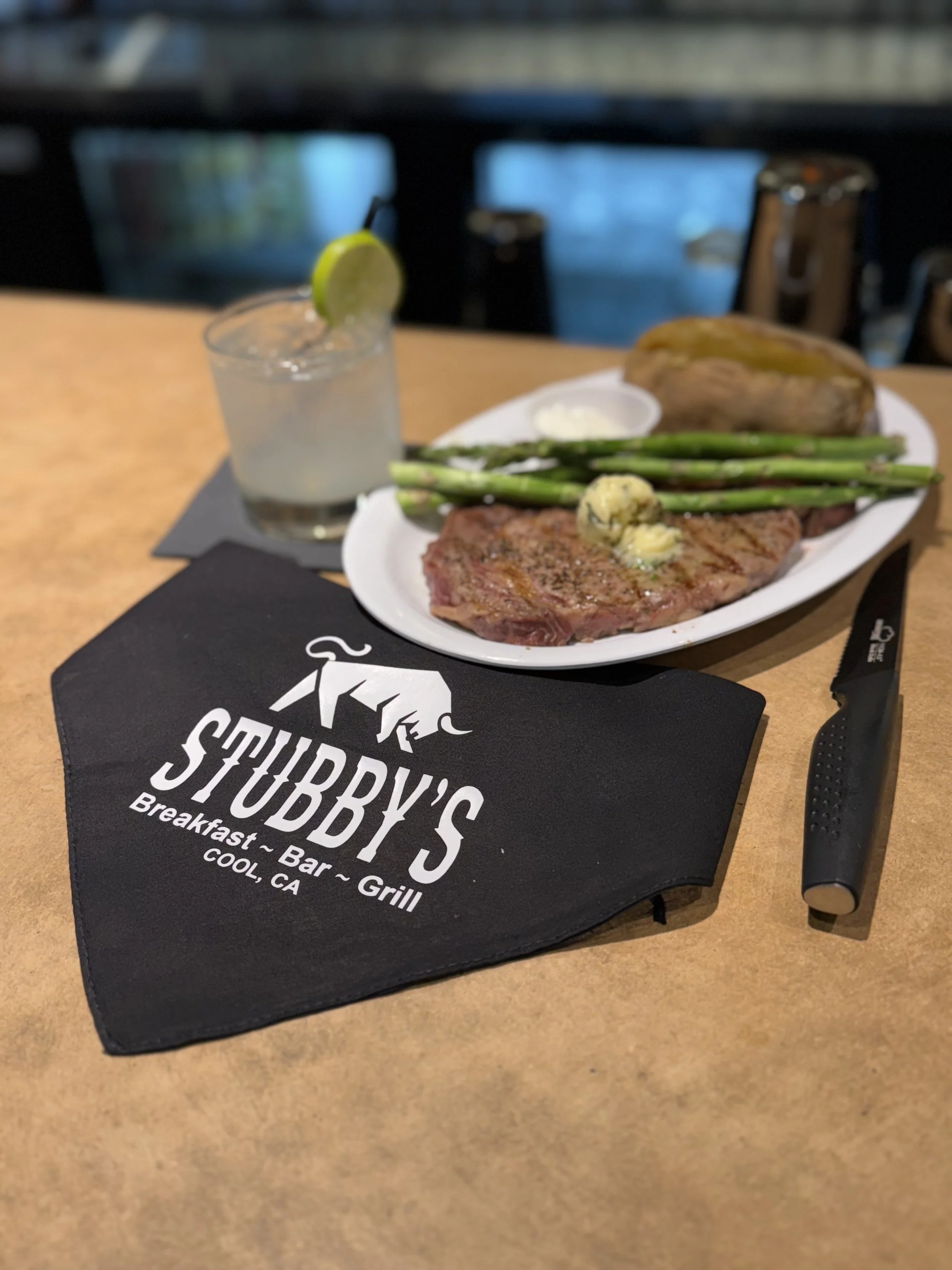 A meal at Stubbby's in Cool, California featuring a glass of lemonade with lime, a plate with asparagus, a baked potato, and a steak topped with butter. A black napkin with the restaurant's logo is on the table, along with a black-handled steak knife