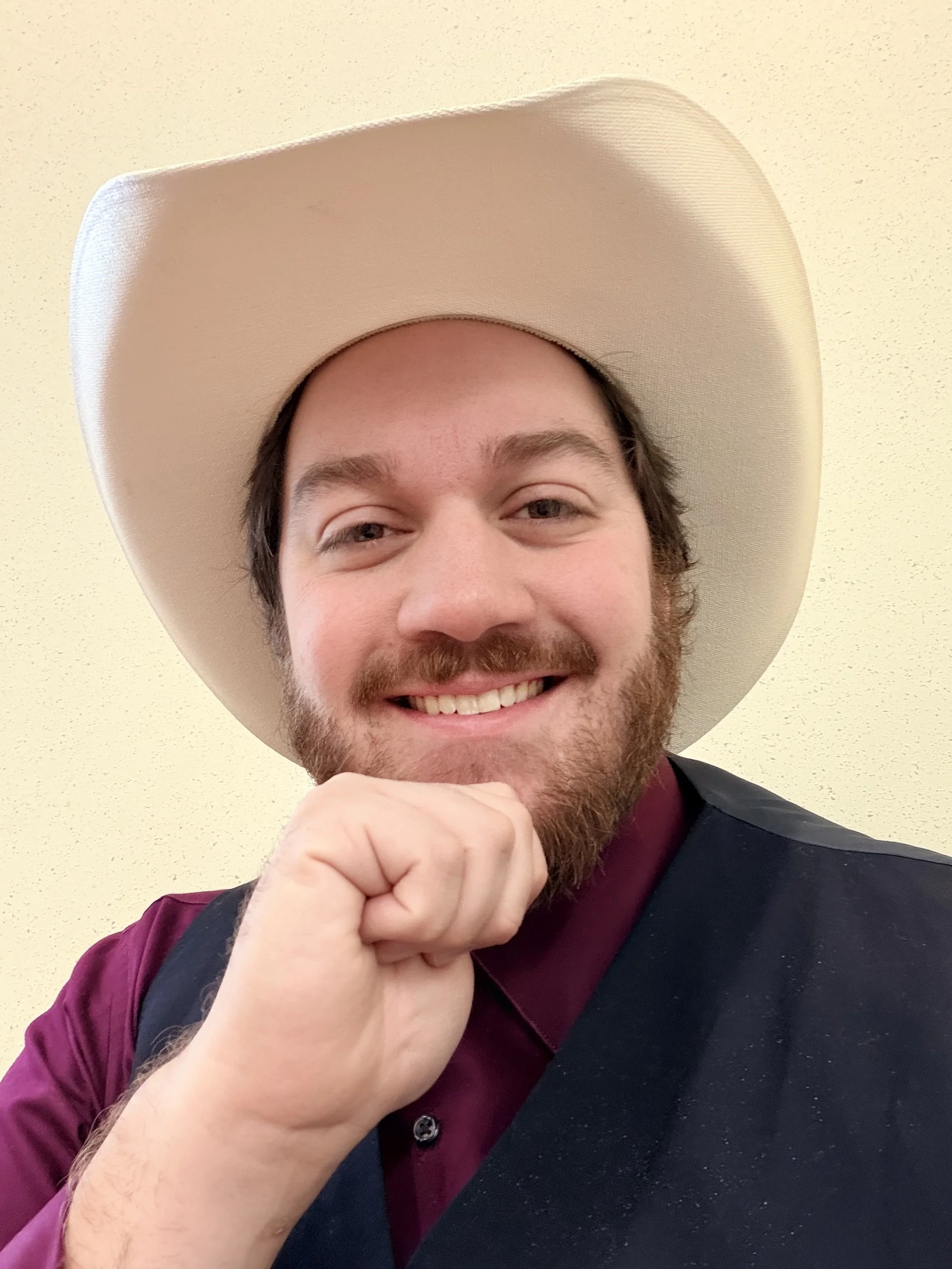 A smiling man with a beard and mustache wearing a large white cowboy hat, a purple shirt, and a dark vest, posing with his fist under his chin against a plain yellow wall.