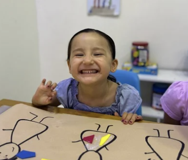 Smiling young girl with dark hair and a purple shirt at a table with paper crafts, in a room with a white wall and colorful background.
