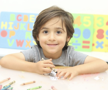 A young boy sitting at a table with crayons, smiling, with colorful alphabet and number posters in the background.