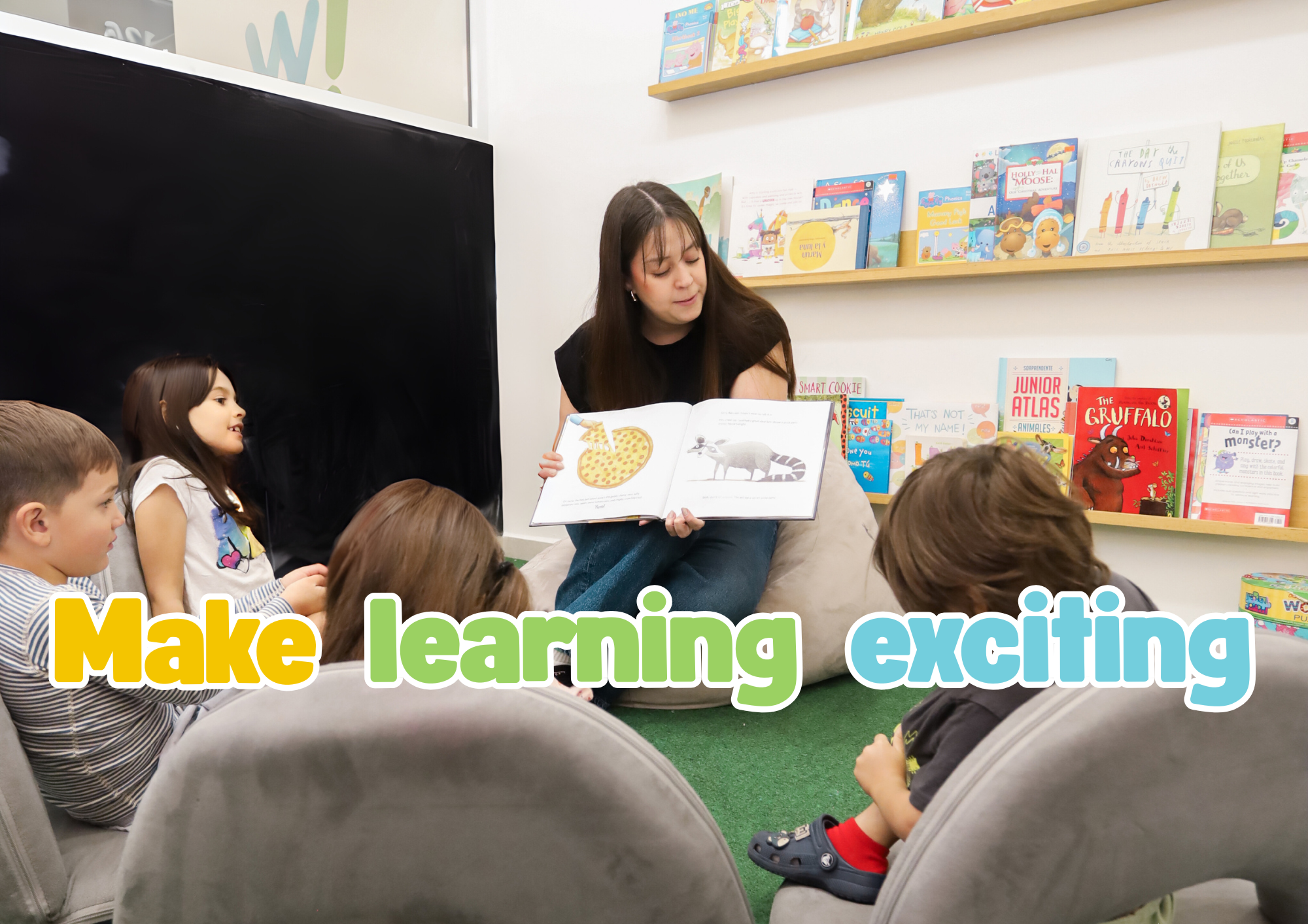 A young woman reading a children's book to a group of children in a library or classroom. The children are sitting on chairs and on the floor, watching attentively. The background has bookshelves filled with colorful children's books. The overlay text says 'Make learning exciting' in bright colors.
