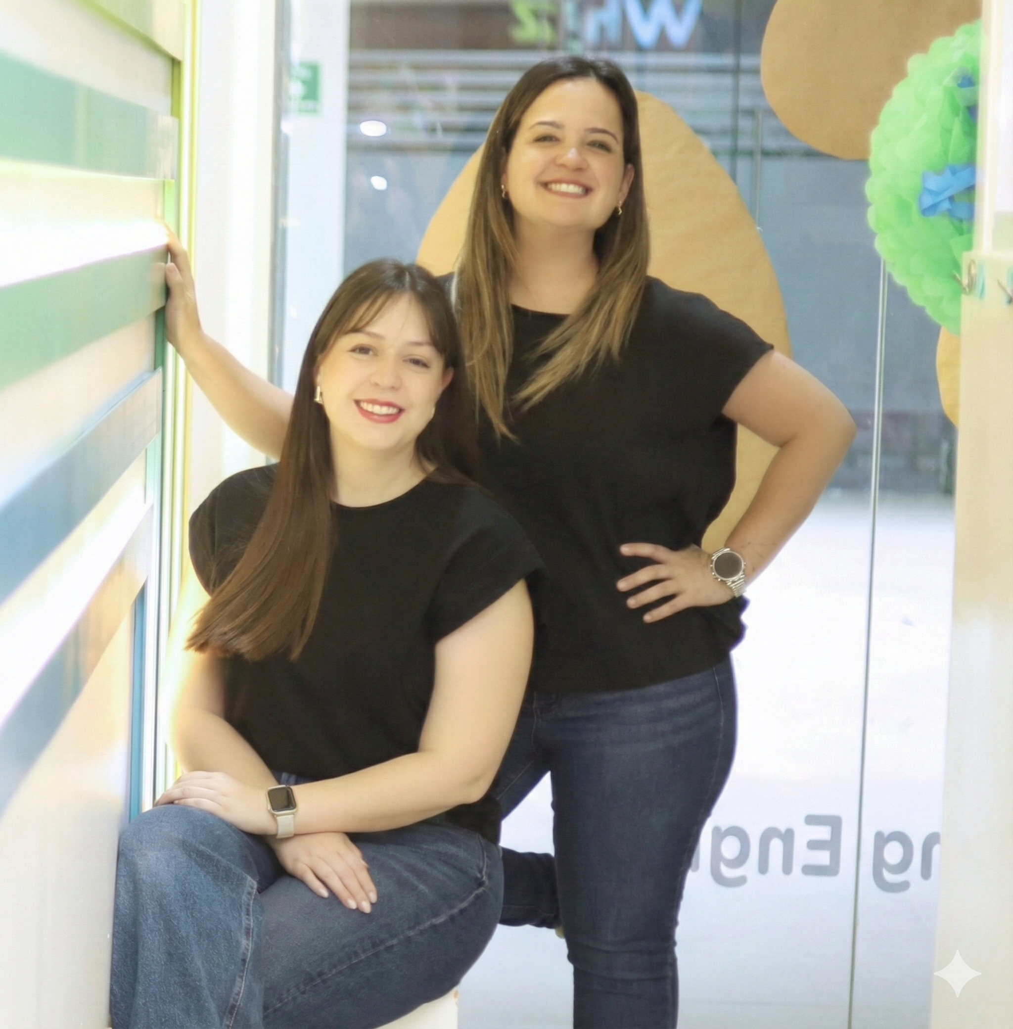 Two women with brown hair, wearing black t-shirts, smiling, posing in front of a glass door with decorations, one is sitting on a ledge and the other is standing.