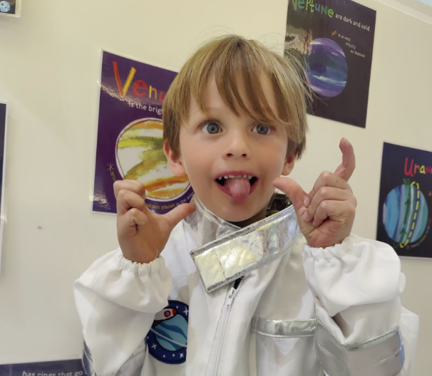 A young boy with brown hair and blue eyes making a silly face, sticking out his tongue, and pointing at his head. He is wearing a shiny silver and white space-themed costume, with posters of planets and space themes on the wall behind him.