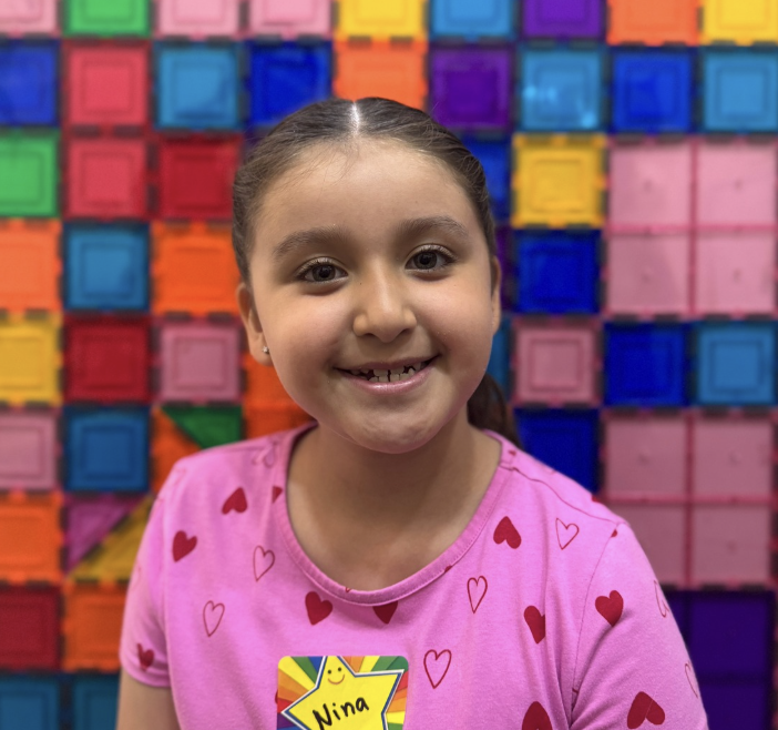 A smiling young girl with brown hair in a ponytail wearing a pink shirt with red and pink hearts, standing in front of a colorful background of stacked books or blocks in bright colors.