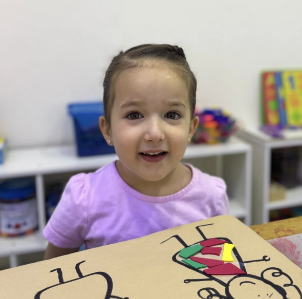 A smiling young girl with a purple shirt sitting at a table in a classroom, with a large drawing of a ladybug made from colored paper in front of her.
