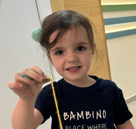 A young girl with a ponytail holding a yellow string or yarn, smiling at the camera indoors.