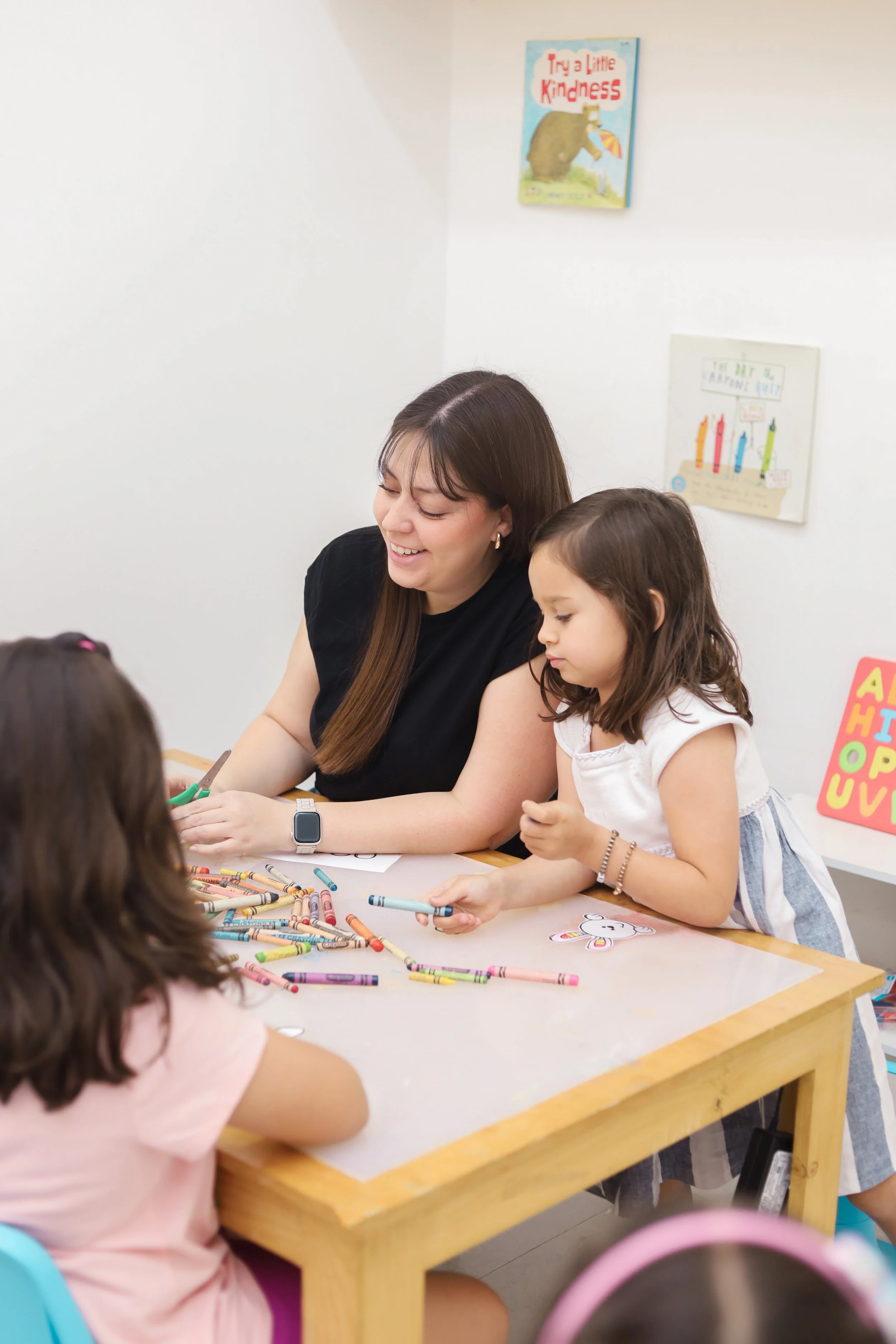 Teacher and young girls engaged in a classroom activity with colorful crayons on a table.