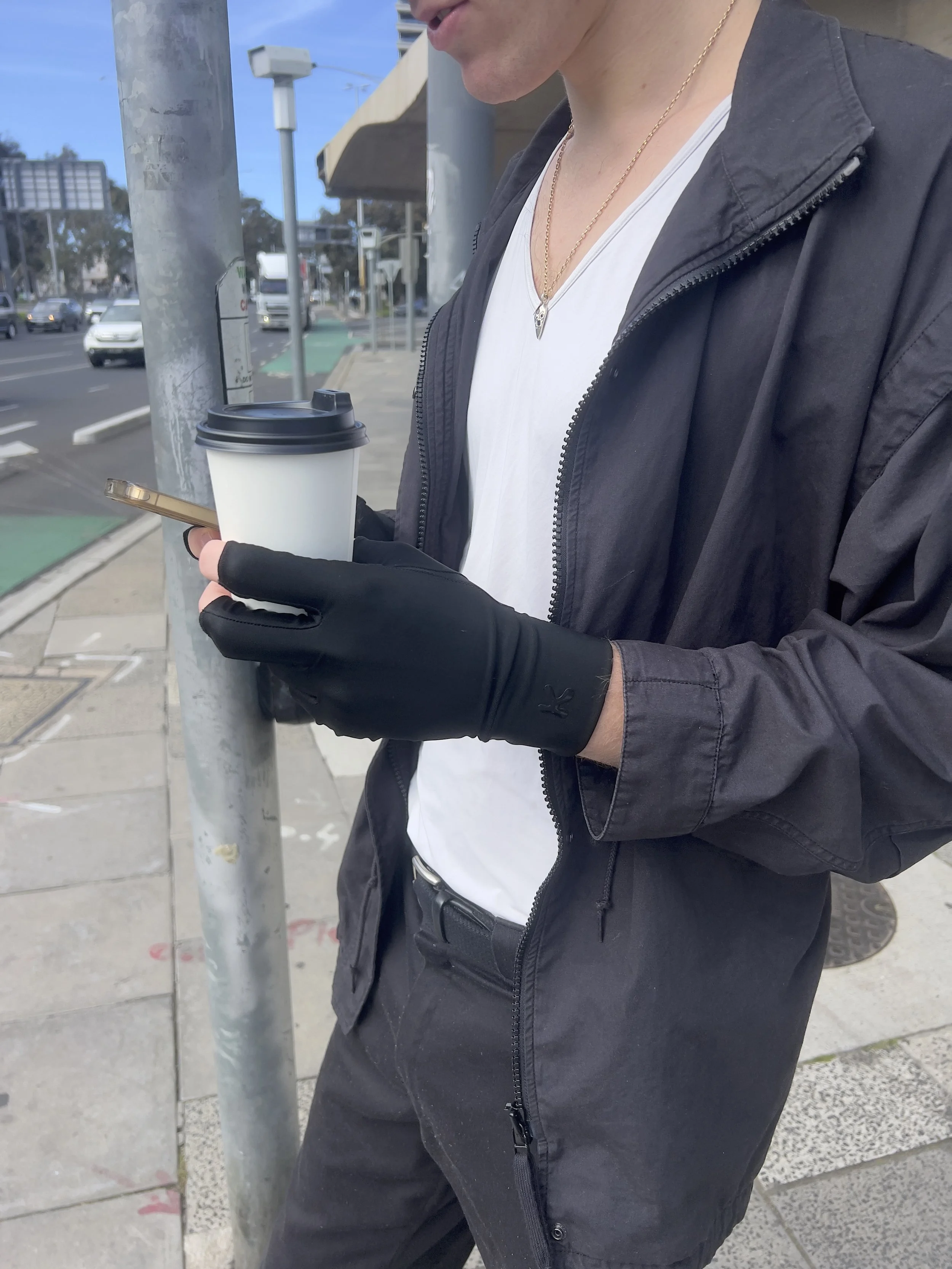 Person wearing black gloves and a black jacket, holding a coffee cup and a smartphone while standing on a city sidewalk with traffic and buildings in the background.