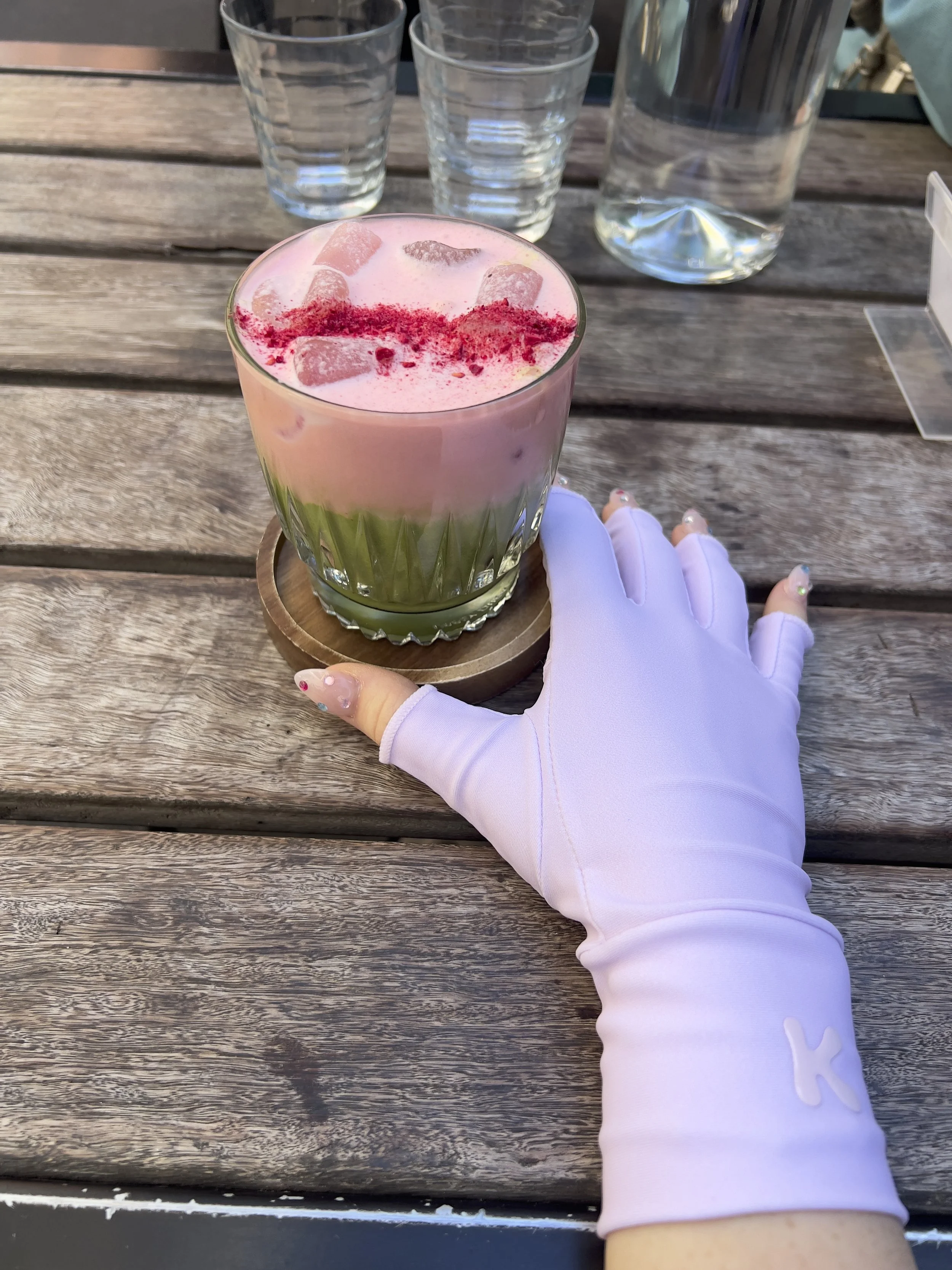 A person's hand wearing a white glove with a 'K' logo resting on a wooden table, holding a drink with pink foam and red powder on top, in a clear glass with ice, surrounded by three empty glasses and a water pitcher.