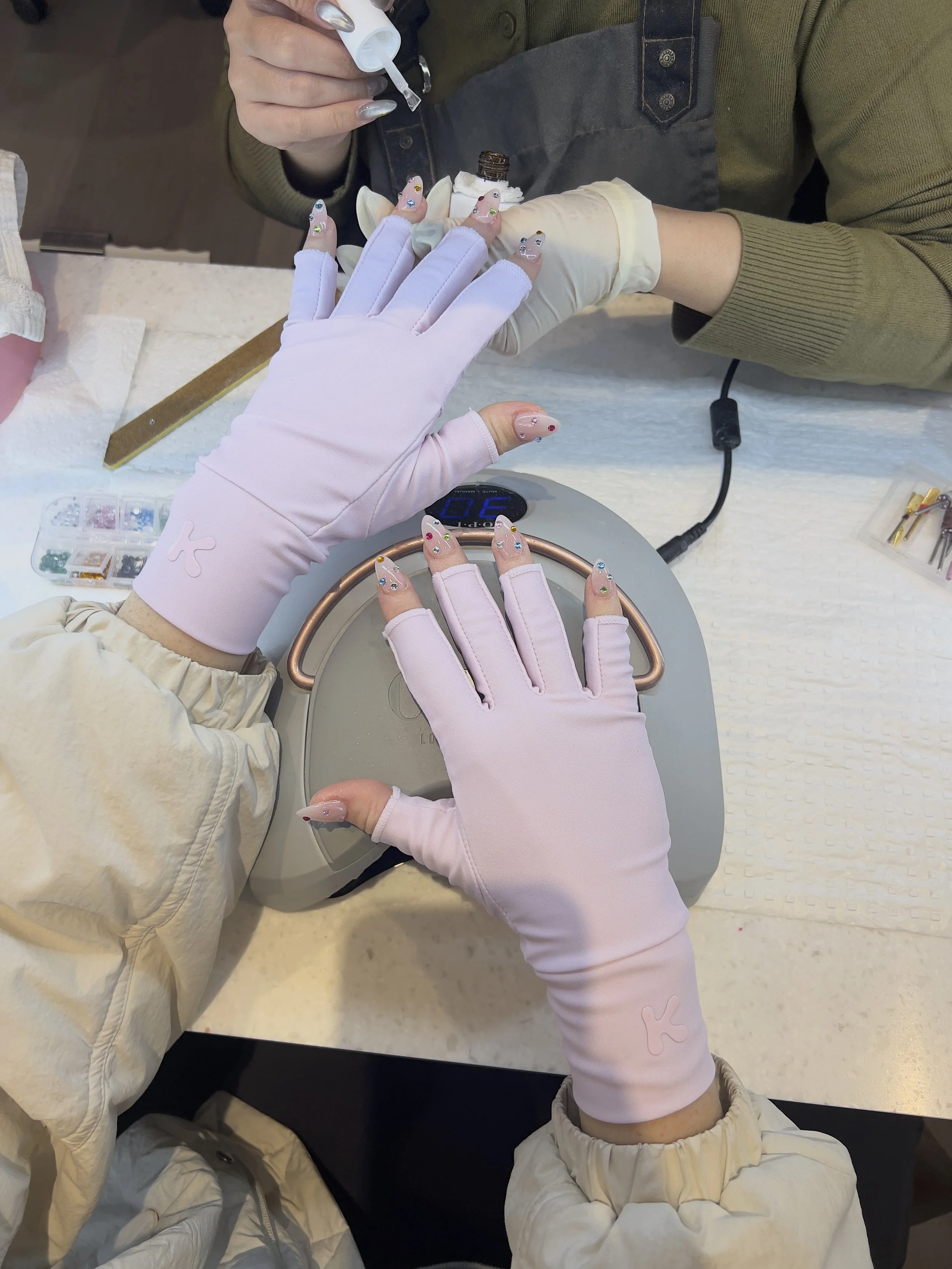 Person receiving a manicure with pink gloves, nail polish, and decorative nail art. Manicure tools and supplies are visible on the table.