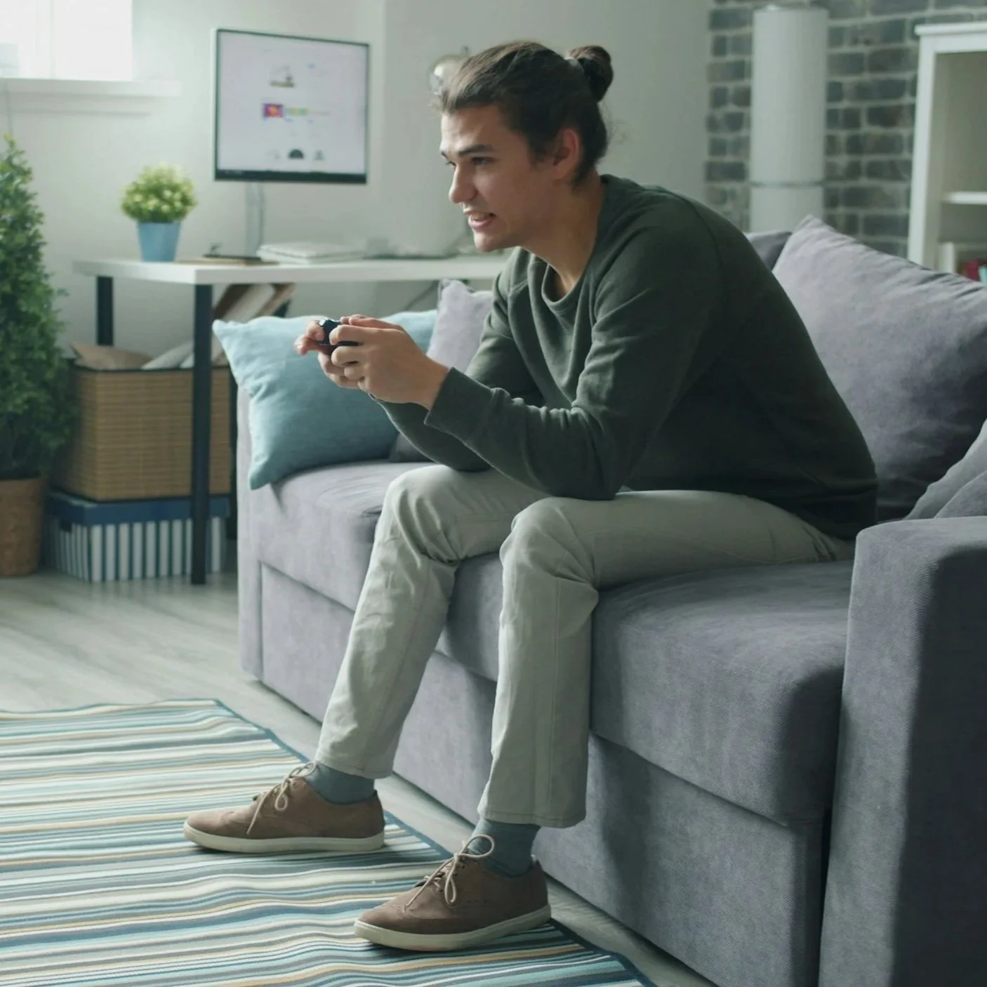 A young man with a ponytail sitting on a gray sofa playing video games, holding a game controller, in a living room with a striped rug and a TV in the background.