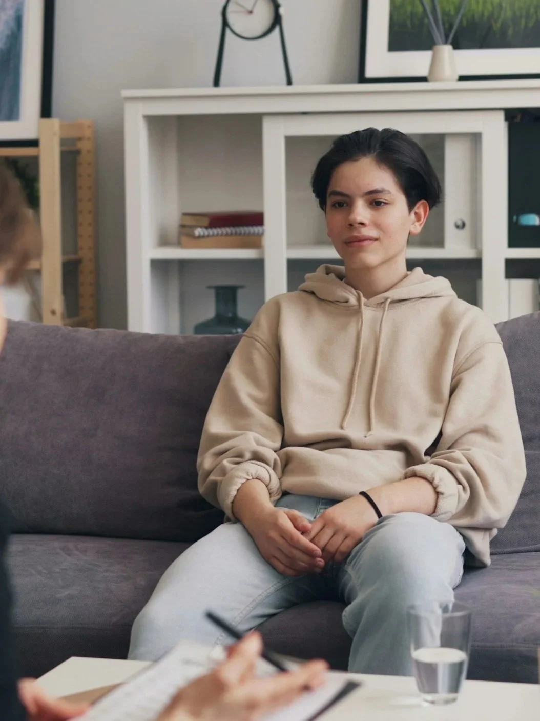 A young person with dark, short hair wearing a beige hoodie and light pants, sitting on a gray couch, talking to someone off-camera in a modern living room.