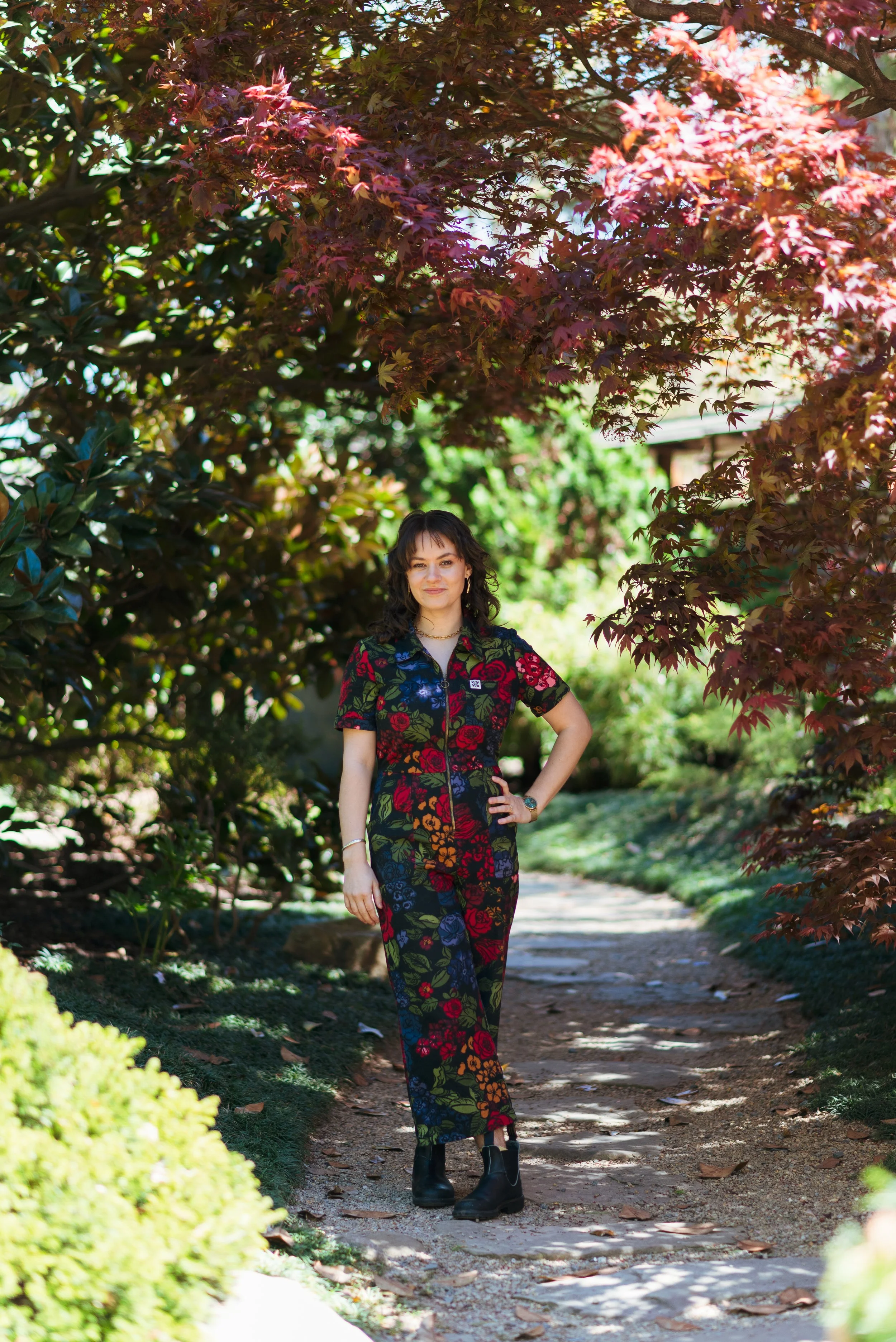 A woman walking on a garden path surrounded by leafy trees and bushes, with sunlight filtering through the foliage.