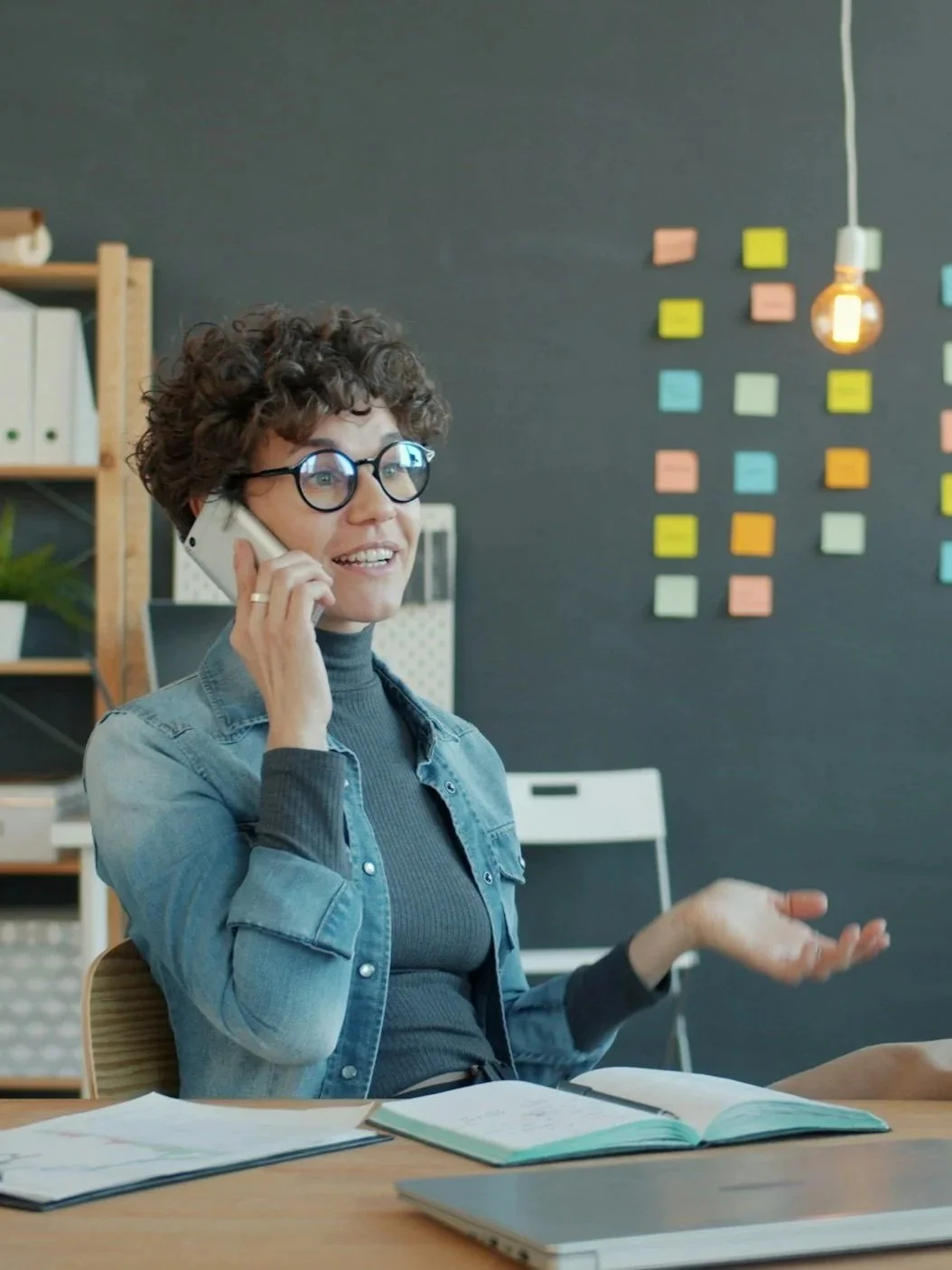 A woman with curly hair and glasses, talking on the phone while gesturing with her hand in a modern office.