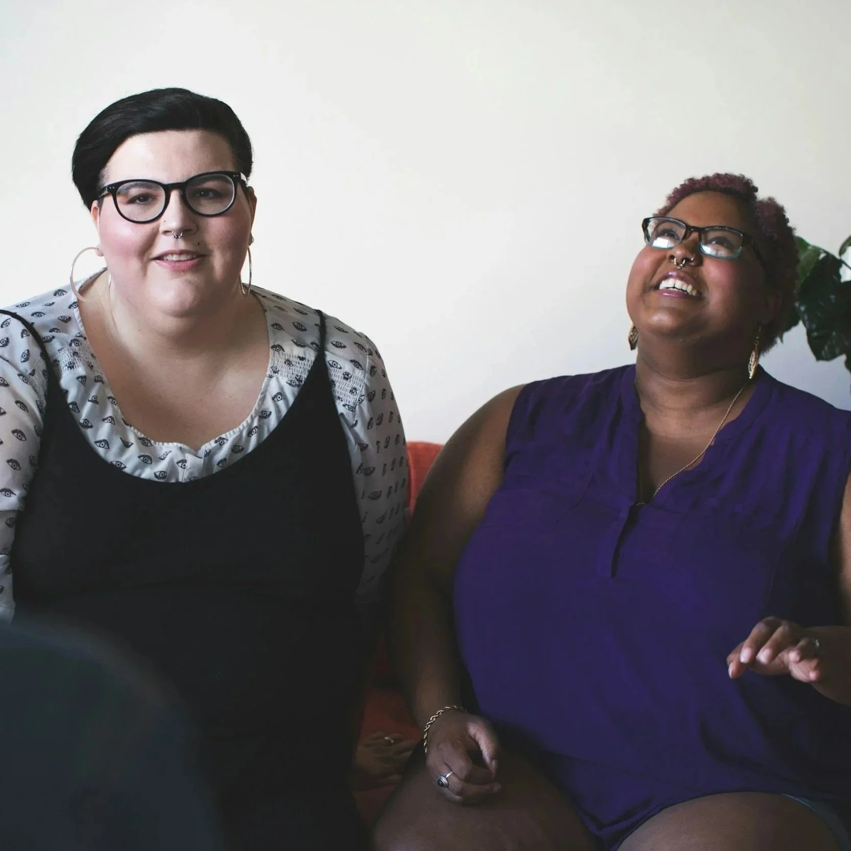 A queer-presenting couple in LGBTQIA therapy sit beside each other on a couch. The person on the right is laughing.