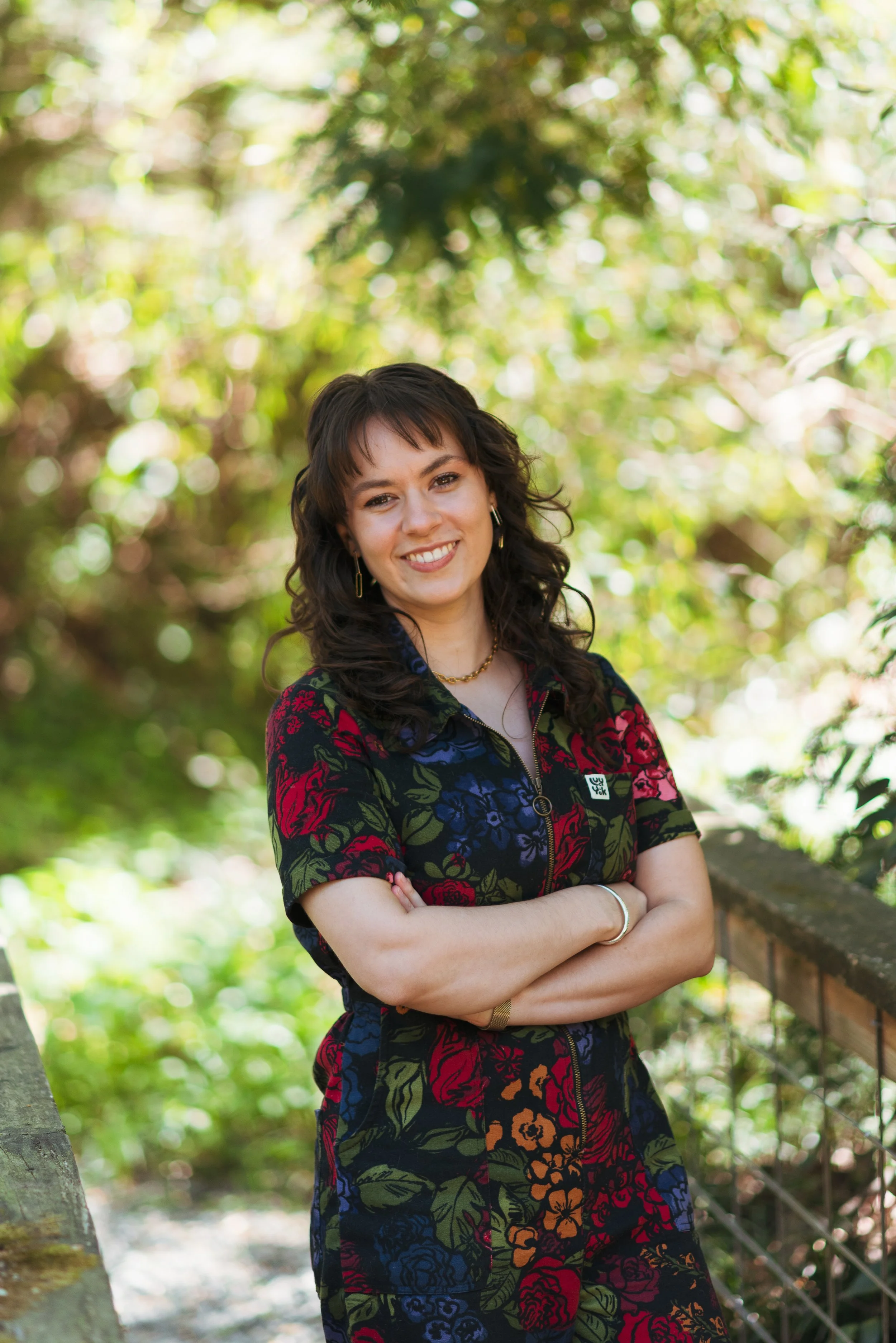 A woman smiling with arms crossed, standing outdoors in a lush green park or garden.