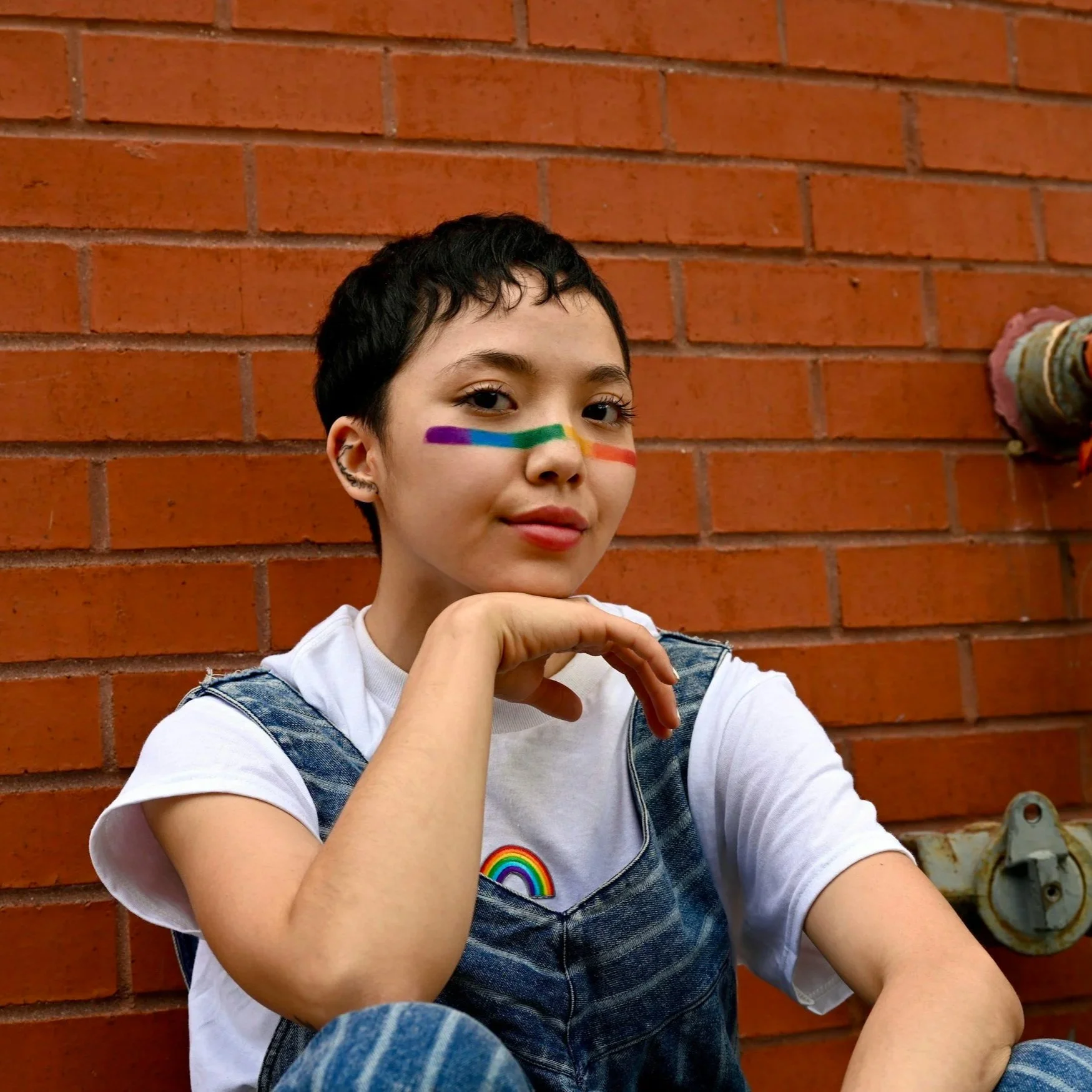 A young person with short dark hair and light skin, sitting against a brick wall. They have rainbow-colored face paint horizontally across their nose and cheeks and a rainbow sticker on their white T-shirt. They are wearing a denim overalls and resting their chin on their hand, with a subtle smile.