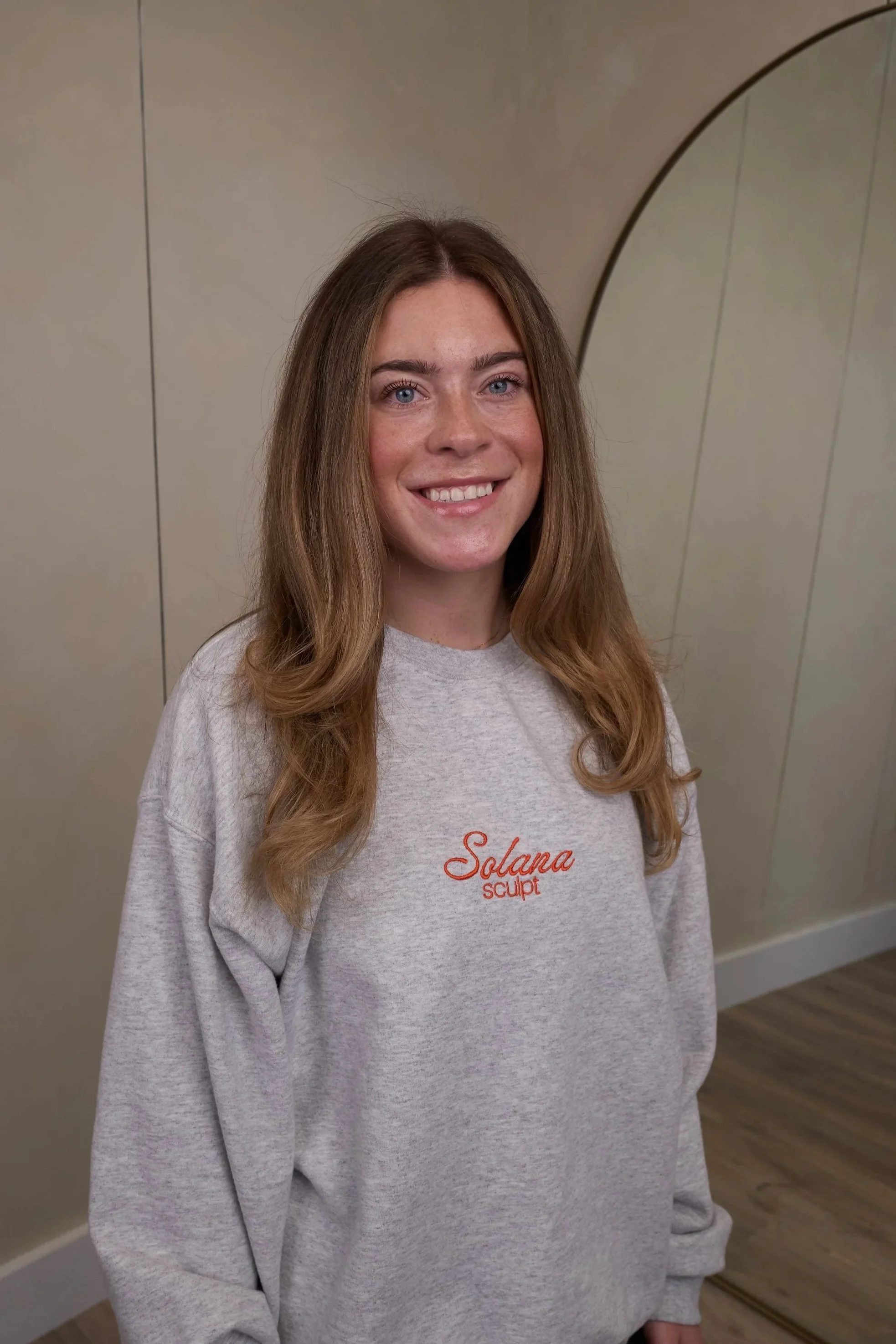 A woman with long, wavy brown hair and blue eyes smiling at the camera, wearing a light gray sweatshirt with red embroidered text that reads 'Solana Sculpt,' standing against a plain light-colored background.