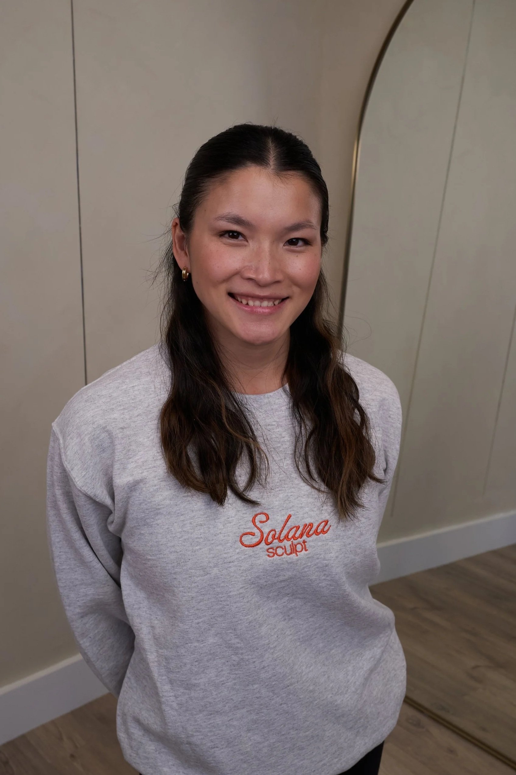 A black dumbbell with a silver handle and a gray rolled-up exercise mat on a light wooden floor, with sunlight casting shadows.