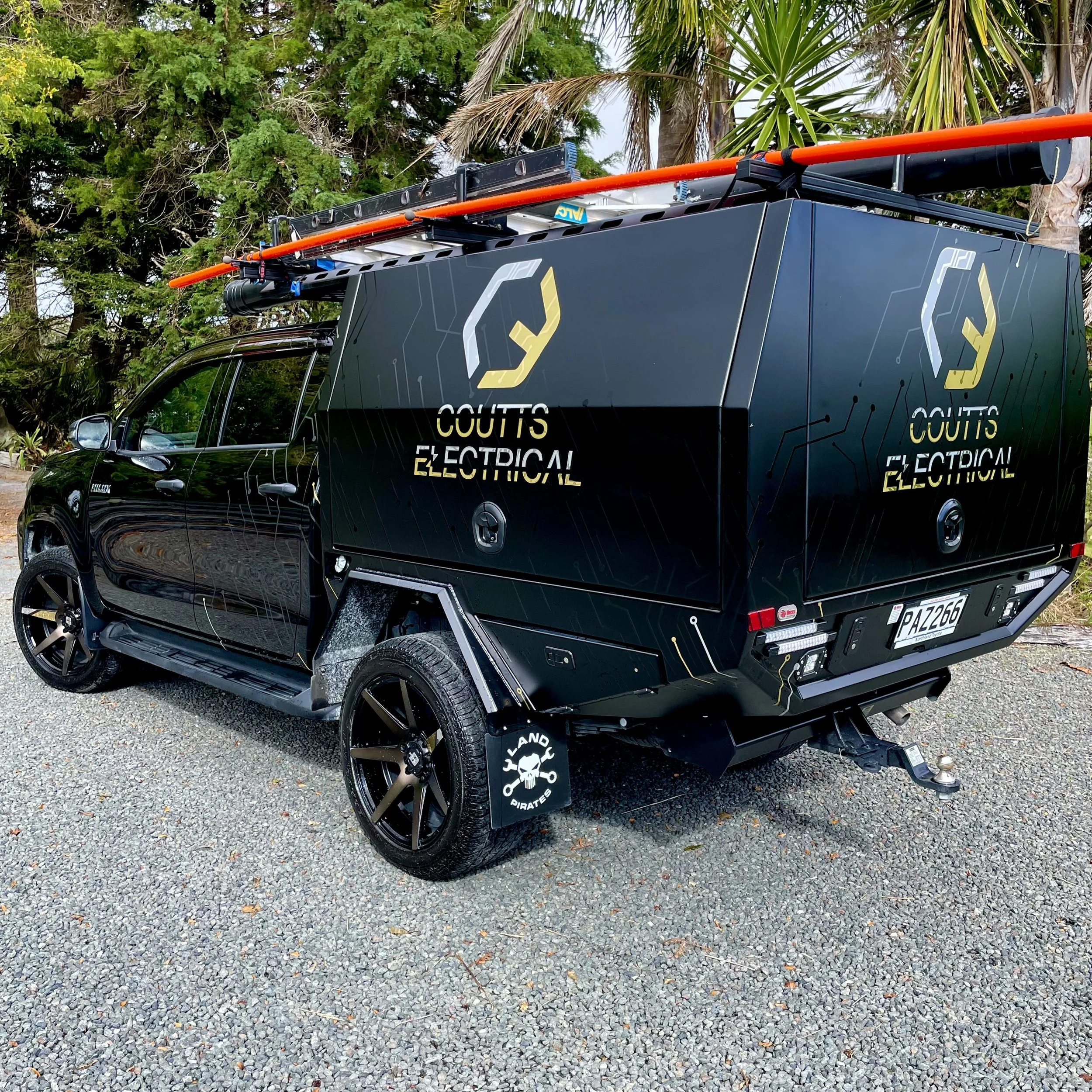 A black electric utility vehicle with specialized cargo box, marked with 'Coutts Electrical' logo and branding, parked on gravel with trees in the background.