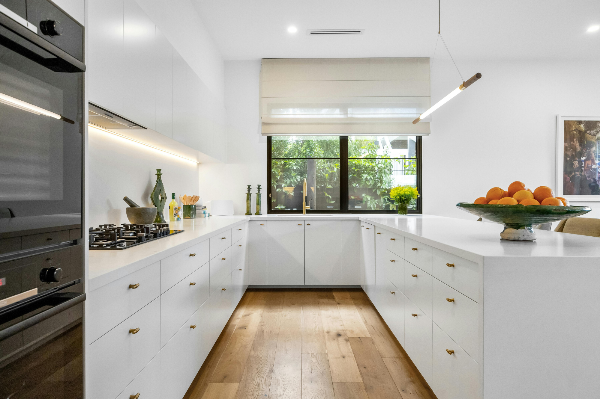 Modern white kitchen with black-framed window, wooden floor, and minimalist decor, including a green bowl of oranges on the counter.